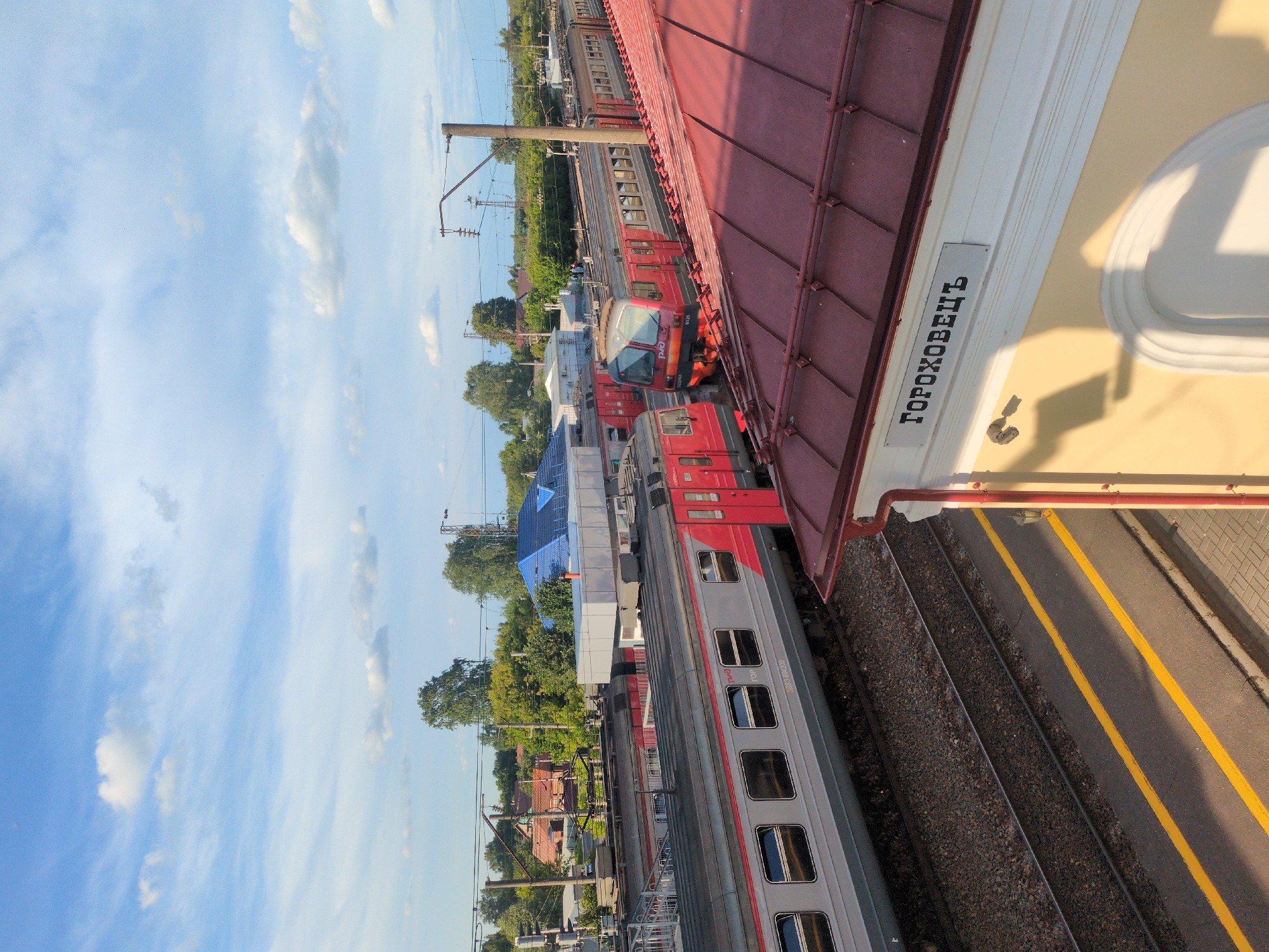 The image captures a view from an elevated position, looking down and across a train station platform. The scene is dominated by a train, predominantly red and grey, parked on the tracks. The train appears to be at rest. To the right of the tracks, a paved platform is visible with two distinct yellow lines painted along its edge. Above the platform, a building with a red-tiled roof and a yellow facade is partially visible. A sign with black lettering in Cyrillic is affixed to the yellow facade. The sky above is a clear blue with scattered white clouds, suggesting fair weather. The lighting indicates it is daytime, likely late afternoon, given the angle of the shadows cast by the building. The surrounding area shows hints of greenery from trees and other structures, possibly indicating a rural or semi-urban train station. The overall impression is one of a typical, operational train station. The visible text on the sign reads "ГОРОХОВЕЦЬ", which is the name of the town, Gorokhovets.