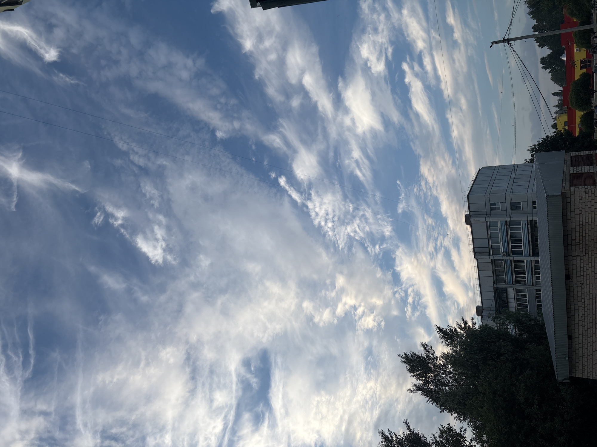 The image captures a view of a cloudy sky above a multi-story building and some trees, likely in a residential area. The sky is a vibrant blue, filled with wispy white clouds that stretch across the frame. Two thin black wires are visible diagonally across the sky, suggesting the presence of overhead power lines. In the lower part of the image, a light blue and gray building with multiple windows is prominent. To the left, a dense dark green tree stands tall. In front of the building, a lower structure with a metallic roof and brick walls is partially visible. The overall scene suggests an outdoor environment, possibly during the daytime, with a mix of natural and urban elements.