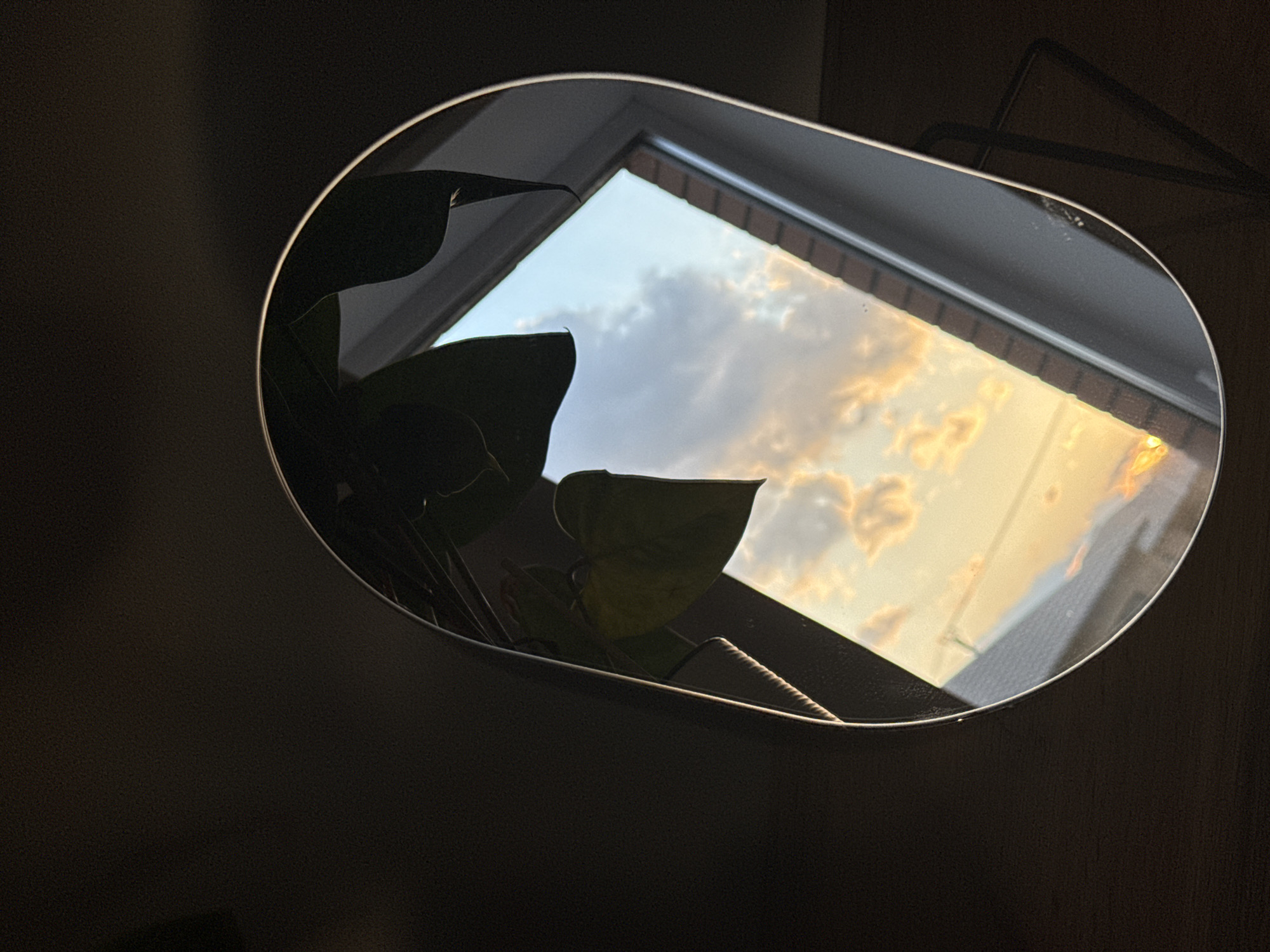 The image shows a reflective, rounded-corner rectangular object (likely a mirror or picture frame) reflecting a window view of a sky with clouds illuminated by the setting sun. The sky is light blue with orange and yellow hues from the sunlight. Plant leaves are partially visible on the left side of the frame, suggesting it is indoors. A dark, blurred background surrounds the reflective object. A metal frame or stand is visible above the rounded-corner object. The image location is unknown and cannot be determined from the information given. Therefore, I am unable to verify whether this image was taken in Chaikovsky, Russia.