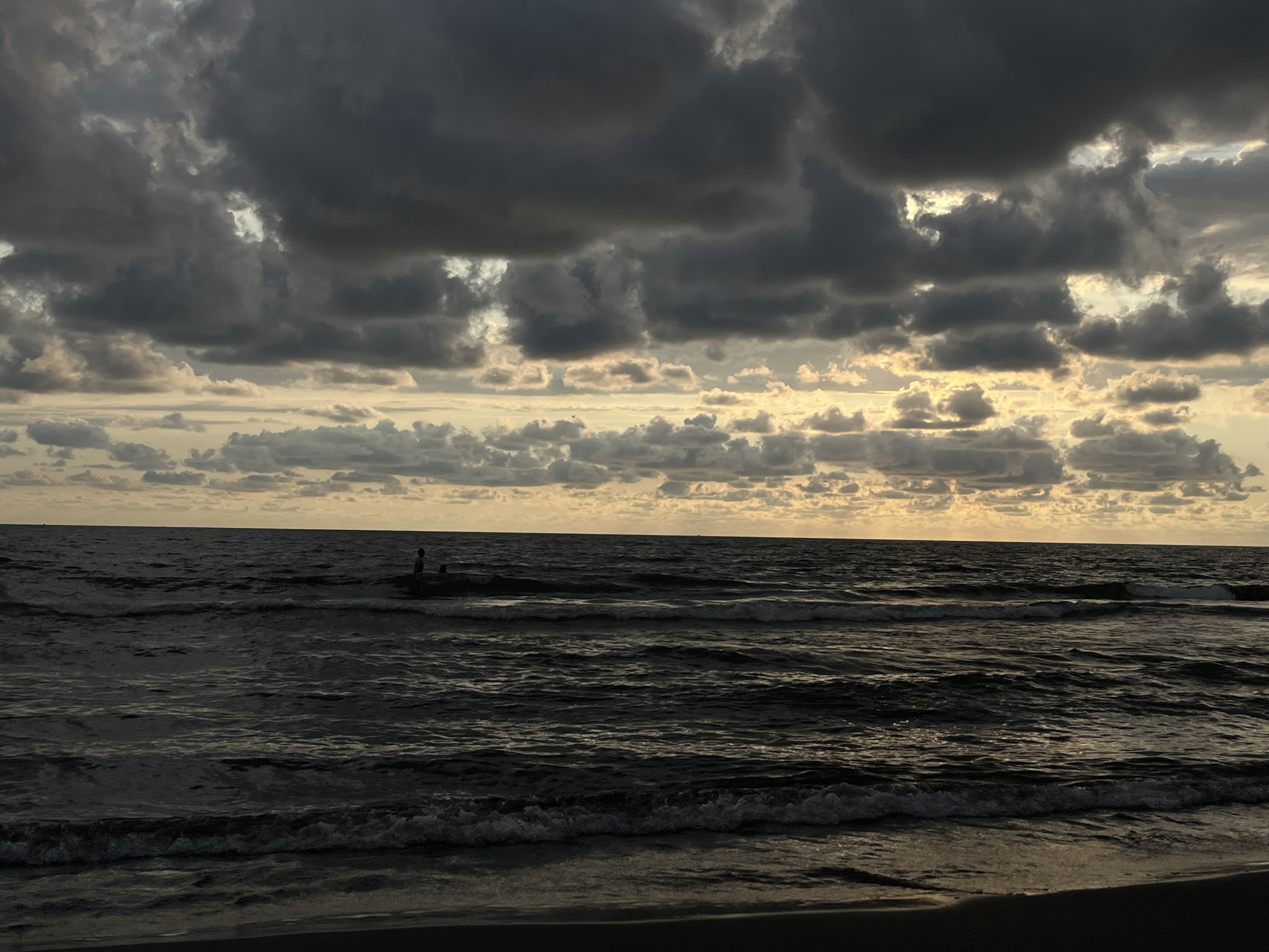 The image captures a serene, yet dramatic, seascape at dusk or dawn, with the sun peeking through heavy, dark clouds. The location is identified as S_upsis T_emi, Georgia. The foreground features the dark sand of a beach, leading to the restless, dark waters of the sea. Gentle waves are breaking on the shore, their crests illuminated by the diffused light. In the mid-ground, two figures, possibly people or animals, are visible in the water, appearing as dark silhouettes against the lighter hues of the sea. The horizon line is clearly defined, separating the tumultuous sea from the sky above. The sky is a spectacle of dramatic cloud formations, with large, dark, ominous clouds dominating the upper portion, while softer, lighter clouds are scattered across the horizon, allowing the warm glow of the setting or rising sun to filter through. The overall mood is one of natural beauty, with a touch of melancholy due to the dark clouds and subdued lighting.