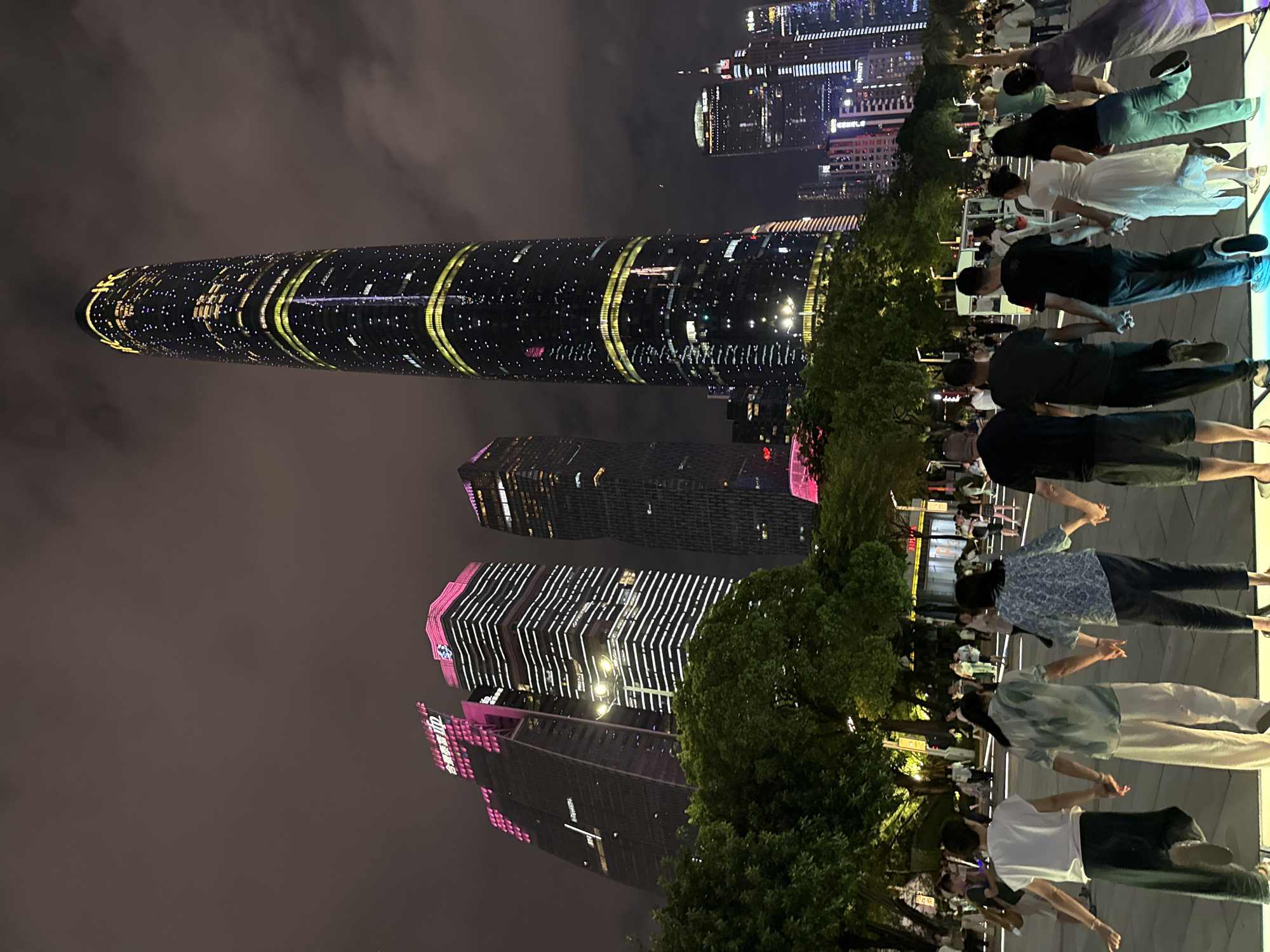 A group of people are holding hands in a line, facing a cityscape at night. The city is illuminated by the lights of numerous skyscrapers, some of which are decorated with colorful LED patterns. The tallest building in the foreground is a cylindrical structure with a distinctive design featuring yellow and white lights wrapping around it. To the right of the cylindrical building, two other skyscrapers are visible, adorned with pink and white light accents. In the background, more buildings create a dense urban skyline. Trees are interspersed throughout the scene, adding a touch of nature to the urban environment. The sky is dark, indicating it is nighttime, and the atmosphere appears slightly hazy. The people are dressed in casual attire, and their posture suggests they are engaged in a shared activity or performance. Some individuals are smiling, conveying a sense of joy or celebration. The overall impression is one of a vibrant urban setting at night, with a community event or gathering taking place. Visible text on one of the buildings appears to be "everup".