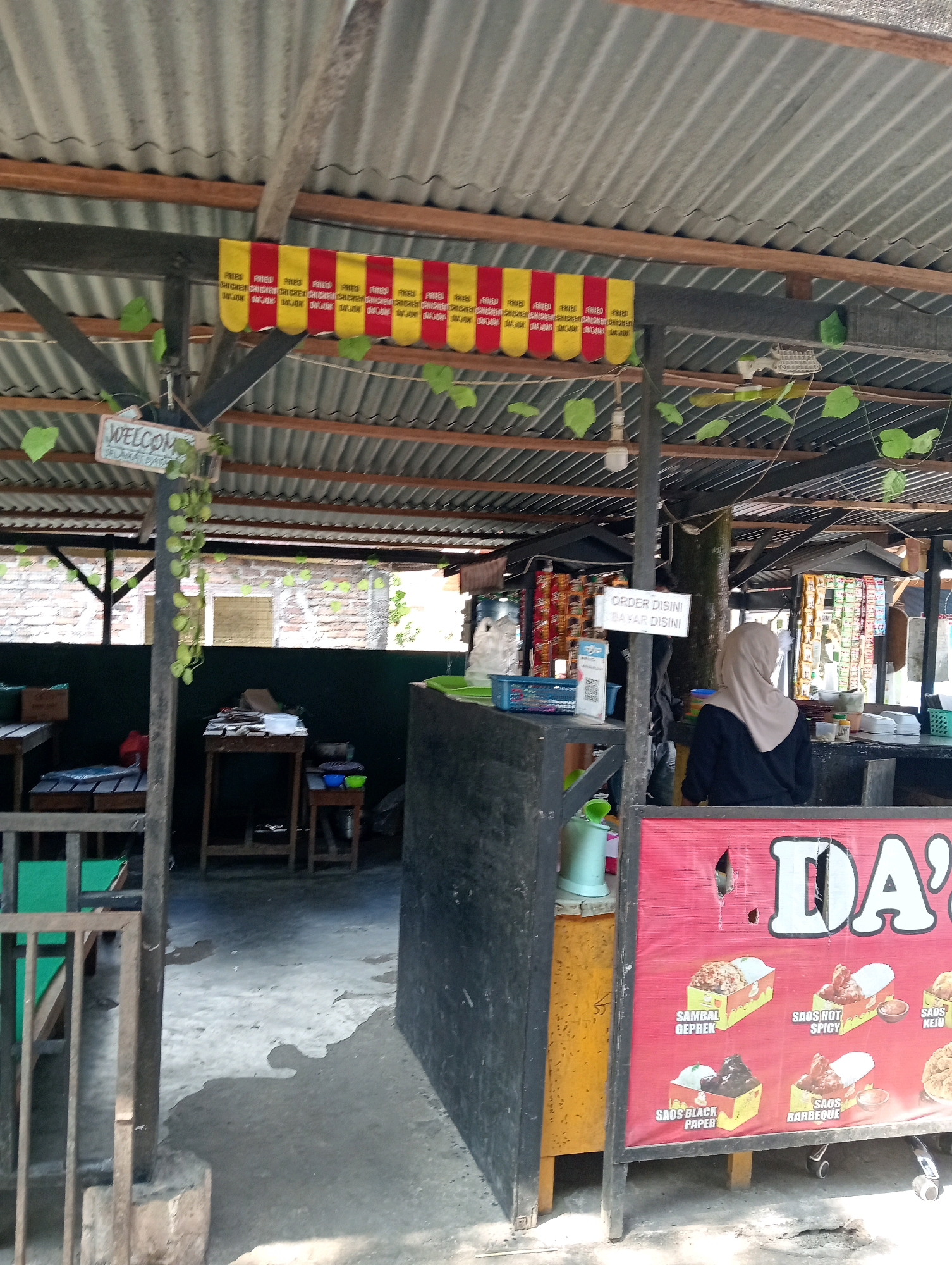 The image depicts an outdoor food stall with a corrugated metal roof supported by wooden beams. A banner with red and yellow alternating squares, each containing the text "FRIED CHICKEN," is hung across the top. Green vines hang from the roof structure. A sign reading "WELCOME SELAMAT DATANG" is visible.
In the foreground, a red banner with the white text "DA'S" is displayed, featuring illustrations of food items such as "SAMBAL GEPREK," "SAOS HOT SPICY," "SAOS KEJU," "SAOS BLACK PAPER," and "SAOS BARBEQUE." A woman wearing a beige hijab and a black top stands behind a counter, facing away from the camera. Various food items and packaging are visible on shelves behind her. To the left of the stall, wooden tables and chairs are arranged. The ground appears to be concrete, with some water stains. The background shows a brick wall and more of the stall structure. The scene is likely a local eatery in Campago, Indonesia, given the Indonesian text on the signs.