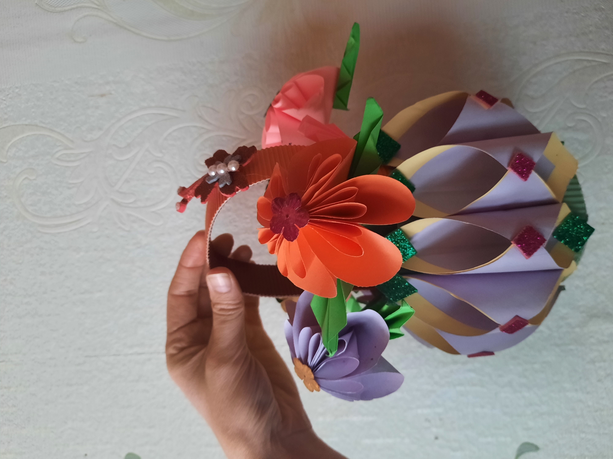 A hand holds a craft project, likely a headdress or decoration. The project is made of paper in various colors, including orange, purple, pink, green, and beige. There are paper flowers and a layered, spherical shape with glittery green and red accents. The background is a white surface with a textured pattern. This image was taken in Архангельское, Russia.