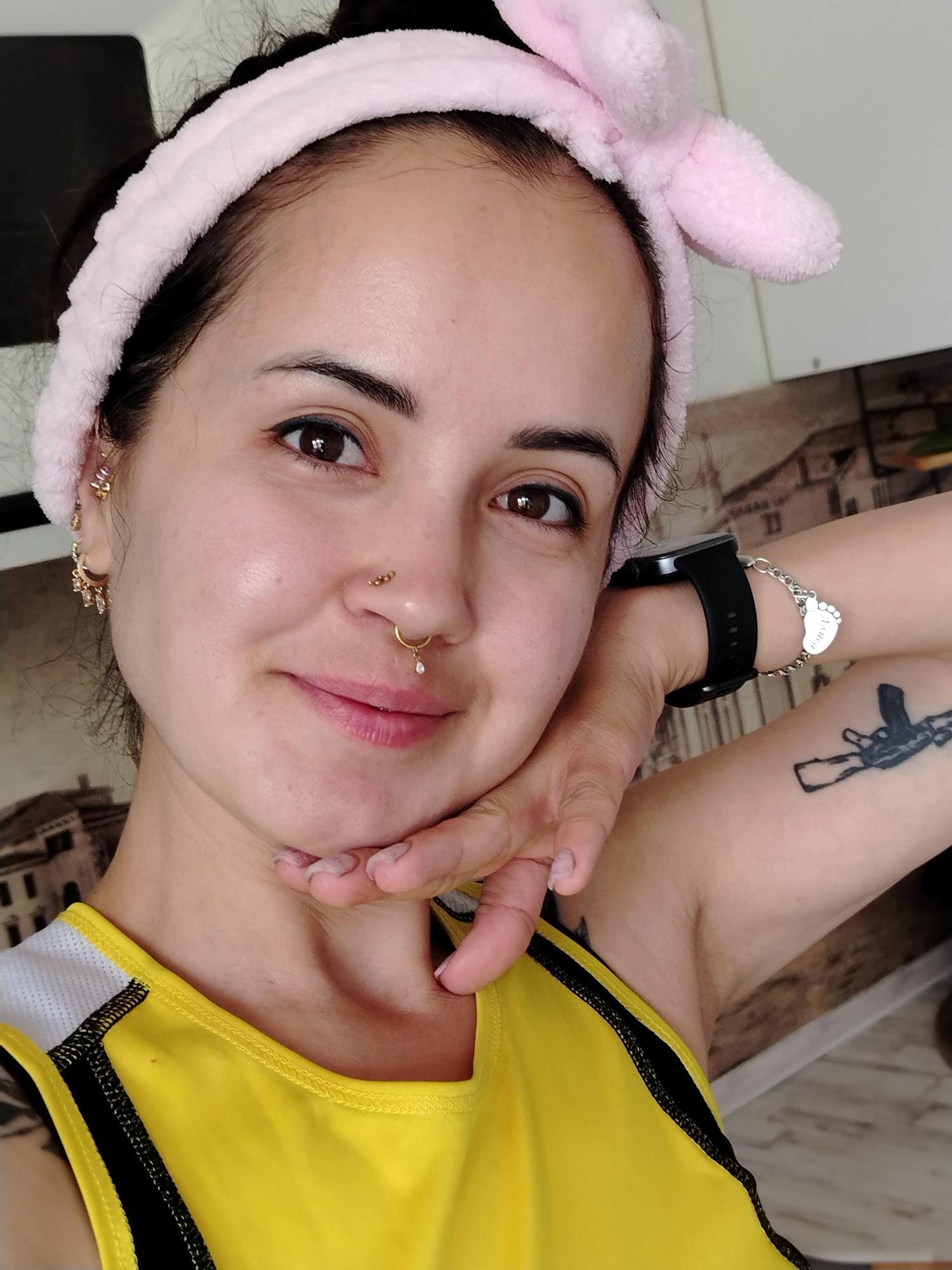 The image displays an adult female in a close-up, chest-up portrait, located in the city of Noviy, Russia. She has dark hair pulled back and wears a pink, plush headband with prominent bunny ears. Her dark brown eyes are directed at the camera, and she exhibits a slight, closed-mouth smile. She has a gold stud piercing with three beads on her right nostril and a gold septum ring featuring a single clear drop-shaped gem. Multiple gold-colored earrings and cuffs are visible on her left ear, including a crescent moon design with hanging clear gems. She wears a yellow athletic tank top with white and black mesh detailing on the left shoulder. On her left wrist, she has a black digital smartwatch and a silver chain bracelet with a heart-shaped charm and a foot-shaped charm inscribed with "Amica." A black tattoo depicting a stylized assault rifle held by a hand is visible on her upper left arm. Her right hand rests gently beneath her chin. The background is out of focus, revealing an indoor domestic setting with a white surface on the upper right and a brownish-toned wall element featuring architectural patterns across the left and central areas.