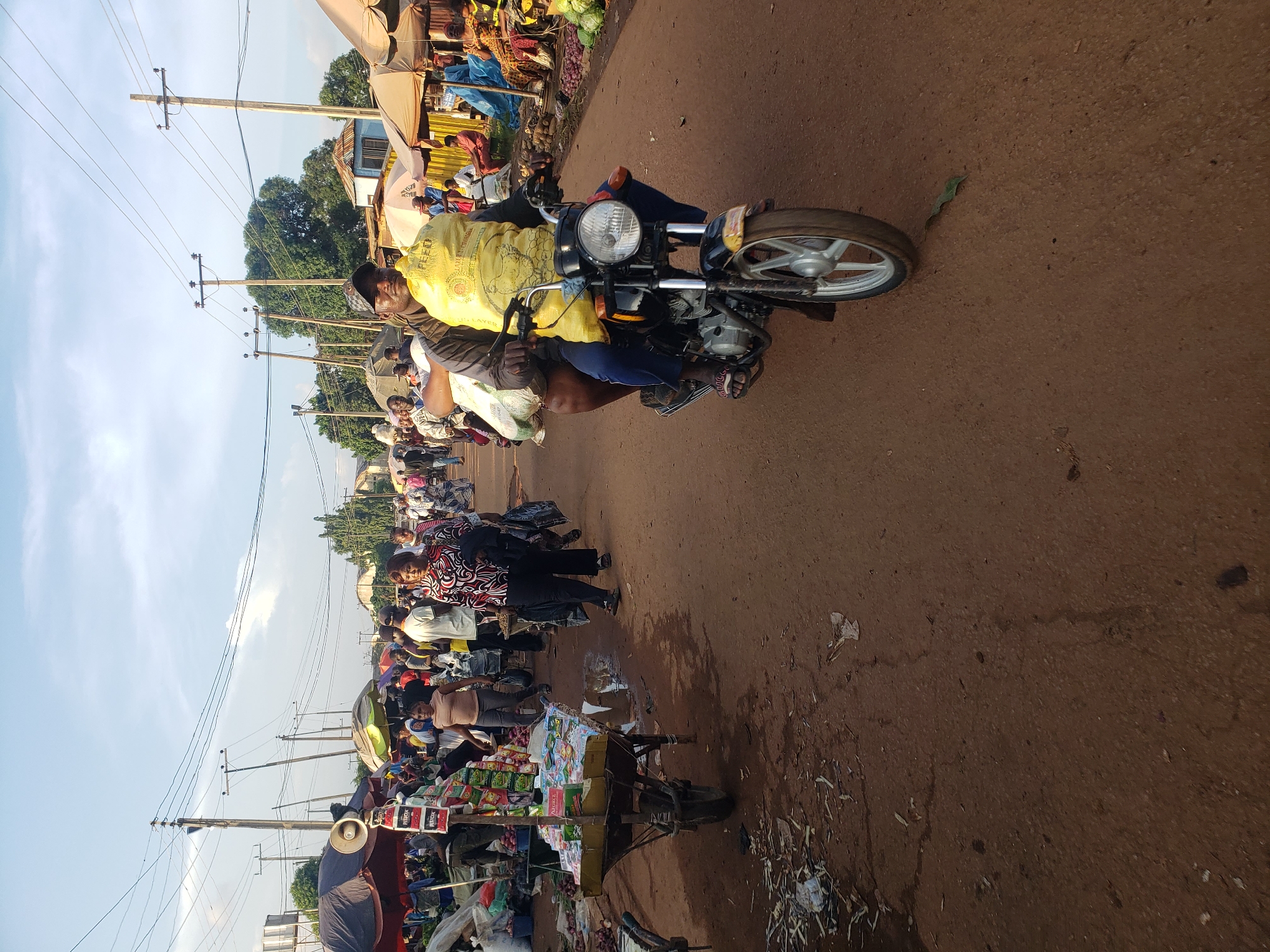 The image depicts a busy street scene in the city of Kakuri Gwari, Nigeria, likely a market area. In the foreground, two individuals are riding on a motorcycle. The person in the front is operating the motorcycle, wearing a dark jacket and a cap. They are carrying a large, bright yellow bag of what appears to be grain or feed on their back. Behind them, another person is seated, also carrying a large bag. The motorcycle is black with silver accents and has a front wheel visible.
The street is unpaved, with a reddish-brown dirt surface, showing some signs of moisture and debris. To the left of the motorcycle, a wheelbarrow filled with items, possibly produce or goods for sale, is parked. Various stalls and umbrellas are set up along the sides of the street, suggesting a marketplace. Several other people are walking in the background, engaged in market activities. Umbrellas of various colors, including red, black, and green, are visible, providing shade. Power lines and utility poles are present in the background, indicating urban infrastructure. The sky above is a light blue with some clouds, and trees are visible in the distance. The overall impression is one of a vibrant, active community hub.