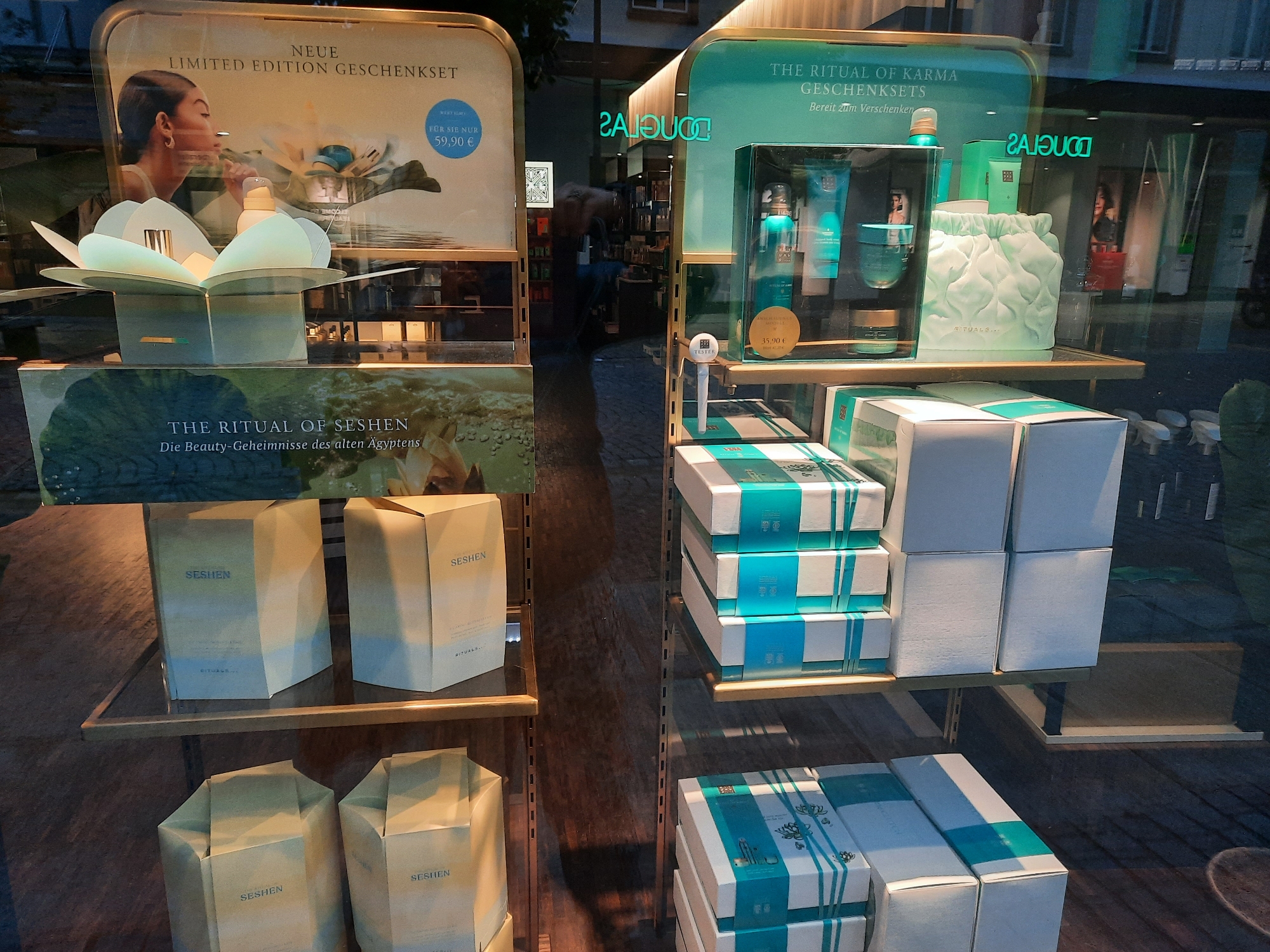 This is a retail window display at a Douglas cosmetics store in Rosenheim, Germany. The display features several gift sets from the "The Ritual of Karma" and "The Ritual of Seshen" lines by Rituals.
On the left side, there is a large display showcasing "NEUE LIMITED EDITION GESCHENKSET" with a price of "FÜR SIE NUR 59,90 €". This set includes a floral-shaped product holder and appears to be a beauty gift set. Below this, there are two stacked gift boxes with the "SESCHEN" branding in beige packaging.
On the right side, the display highlights "THE RITUAL OF KARMA GESCHENKSETS Bereit zum Verschenken". This section features several teal-colored beauty products in a clear display case, including bottles and jars, along with a light green quilted pouch. A smaller gift set is priced at "35,90 €". Below this, there are stacks of gift boxes with teal and white striped packaging.
The store interior is visible through the window, showing shelves stocked with various cosmetic products and a reflection of a person in the background. The outdoor environment appears to be nighttime or evening, with artificial lighting illuminating the display.