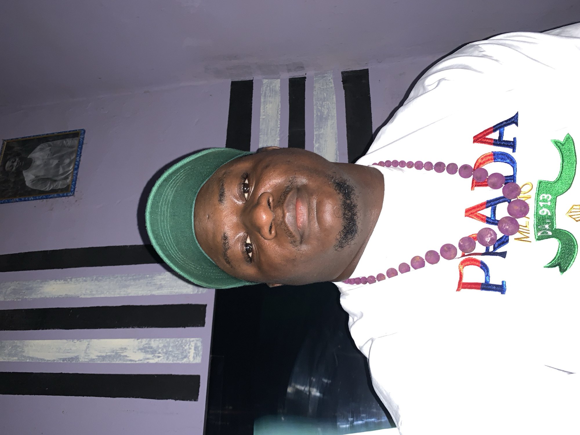 A portrait photograph depicts a dark-skinned male wearing a white t-shirt with red, white, blue, and green lettering, and a green baseball cap. He is also wearing a purple beaded necklace. The background consists of a light purple wall with alternating black and white vertical stripes. In the upper left corner of the image, there is a framed picture hanging on the wall. The photo was taken in Akwanga, Nigeria.
