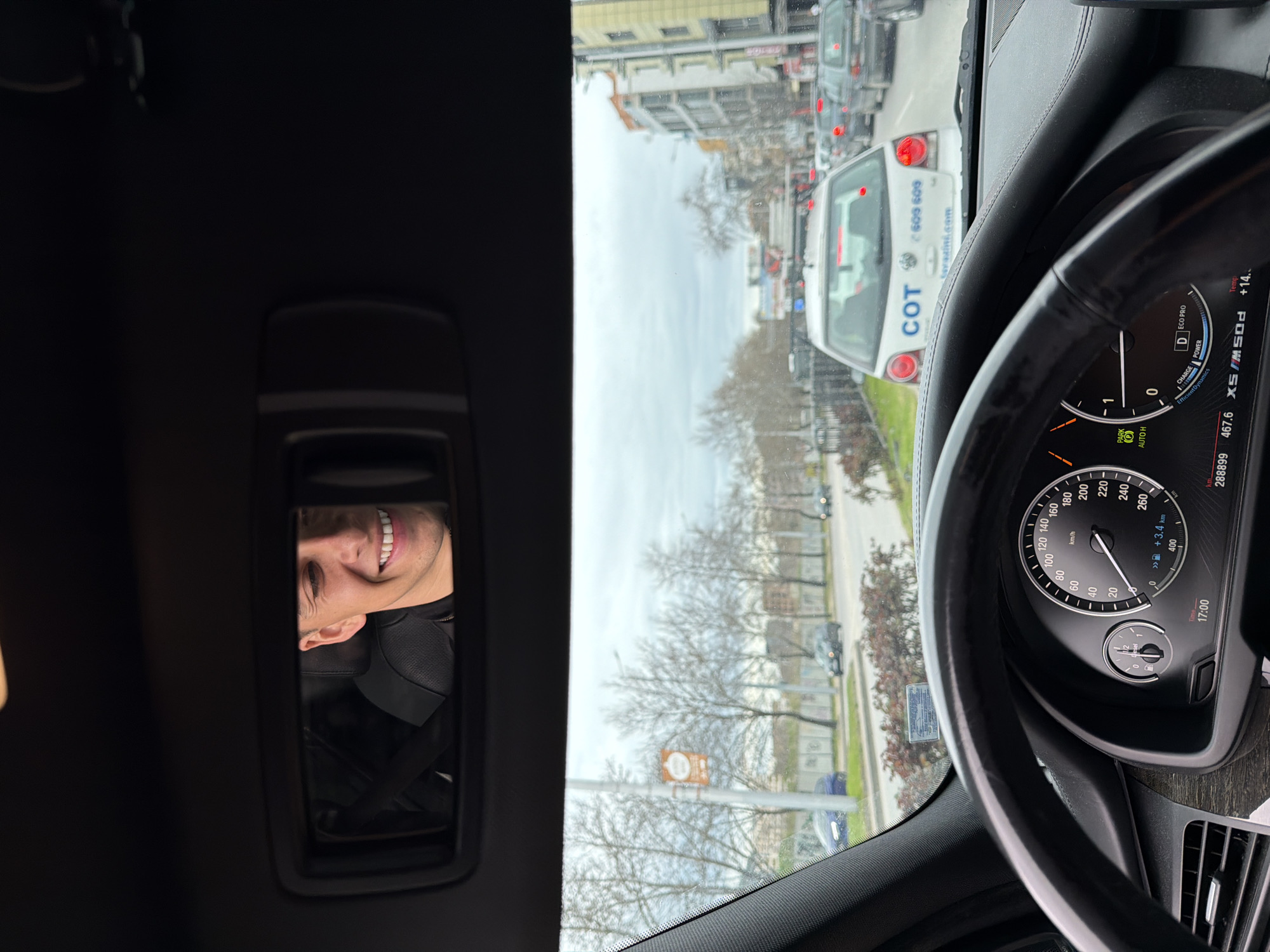 A person is visible in the visor mirror of a car. The car is parked in traffic, with a "COT" branded vehicle (probably a security car) directly in front. The car is in park and the outside temperature is 14 degrees Celsius. The license plate of the car in front is obscured. Through the car's windshield, a streetscape with leafless trees and buildings is visible. The street signs indicate a location in Plovdiv, Bulgaria.