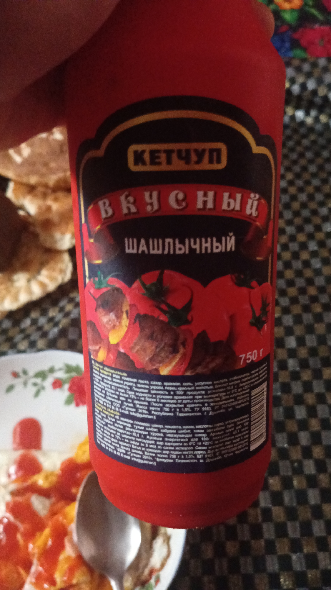 A close-up, slightly blurry shot shows a red bottle of ketchup with a black and yellow label. The label features the word "КЕТЧУП" (Ketchup) in yellow text within a black banner, followed by "ВКУСНЫЙ" (Delicious) in red script. Below this, "ШАШЛЫЧНЫЙ" (Shashlik) is written in white text. The lower part of the label displays an image of grilled meat skewers with red bell peppers and tomatoes. To the right of this image, "750 г" (750g) is printed. The bottle appears to be held by a hand at the top, and is positioned over a plate of food, partially visible at the bottom left, which appears to be eggs with ketchup. The background consists of a checkered cloth. The scene is indoors.