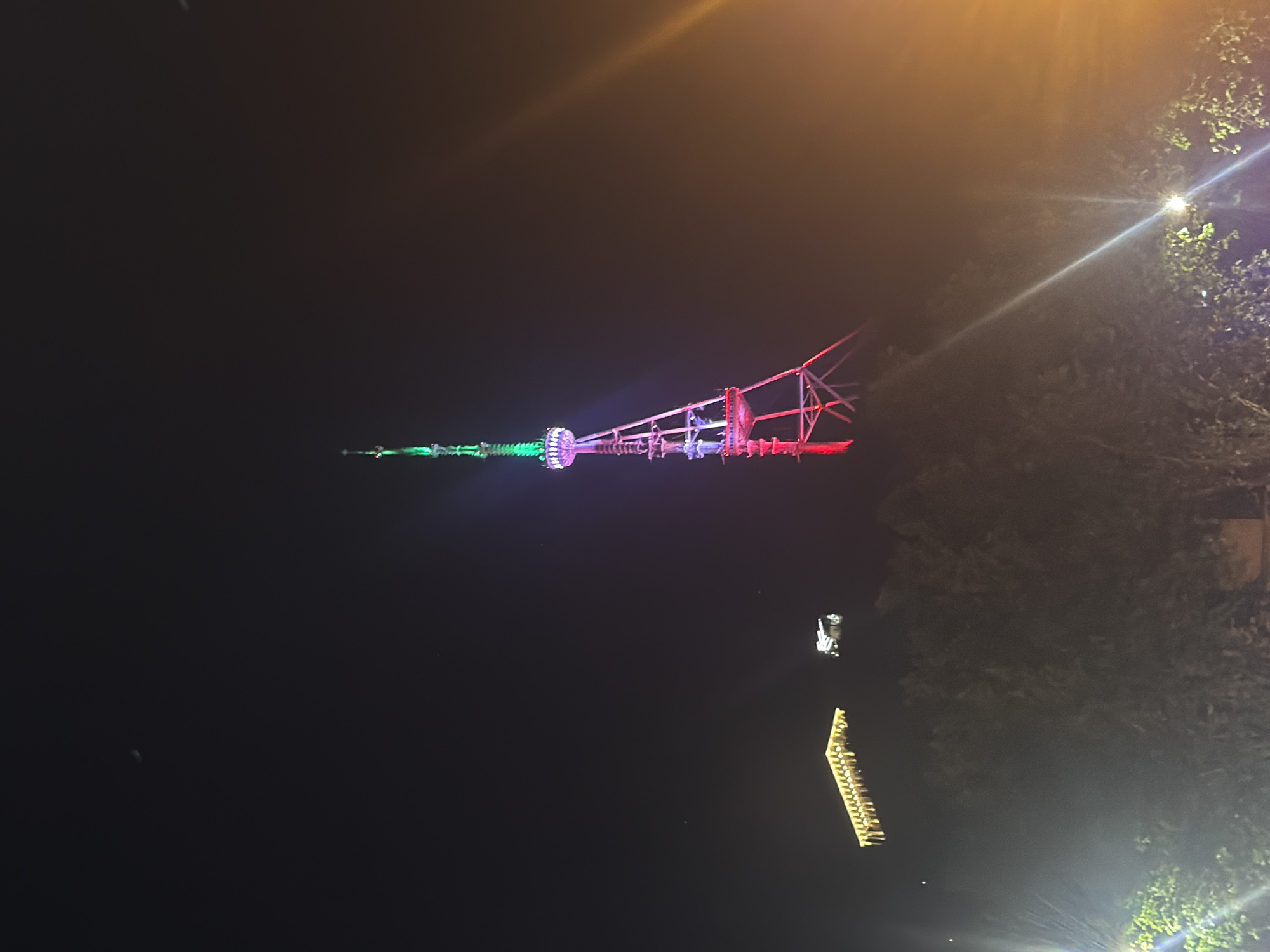 The image displays a night scene featuring a tall, illuminated tower as the central subject. The image appears to be rotated 90 degrees clockwise. When viewed upright, the tower is vertically oriented against a dark night sky.
The tower is brightly lit with various colors. Its slender top section glows green, followed by a wider segment illuminated with green and purple lights. The lower, broader section of the tower, which has a more intricate, lattice-like structure, is lit in red.
To the right of the tower, and slightly in the foreground, are dark trees or dense foliage. Several bright light sources are visible within this foliage, creating lens flares that radiate across parts of the image.
In the lower right portion of the image (when oriented correctly), there are two other illuminated structures. One is a small, box-like structure with bright white lights, resembling a small building or booth. Below and to the left of this, a long, horizontal structure, possibly an elevated walkway or bridge, is brightly outlined with numerous yellow lights.
The scene suggests an outdoor setting at night, possibly within an urban area, park, or amusement park, given the decorative lighting and varied structures. No people or visible text are present. The clear dark sky indicates fair weather.