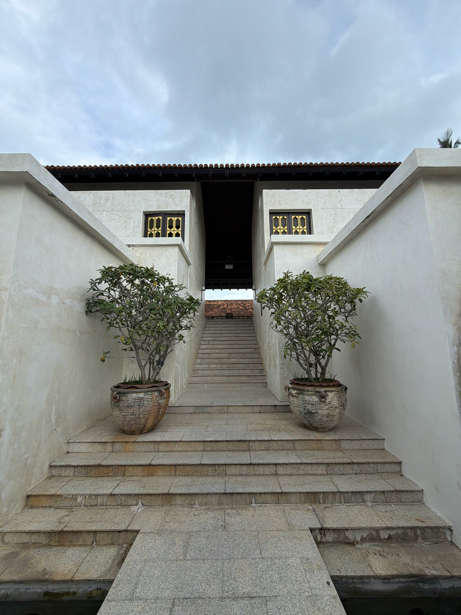 In the city of Ko Samui District, Thailand, a symmetrical outdoor staircase ascends towards an opening in a building. The building has a light-colored facade, a dark tiled roof, and two windows with gold-colored grilles. Flanking the staircase are two large potted plants with lush green foliage. The stairs are constructed of grey stone slabs, with some showing signs of wear and discoloration. A walkway extends from the foreground, leading onto the first few steps of the staircase. The sky above is overcast, with diffused light. The scene depicts a tranquil architectural entrance.