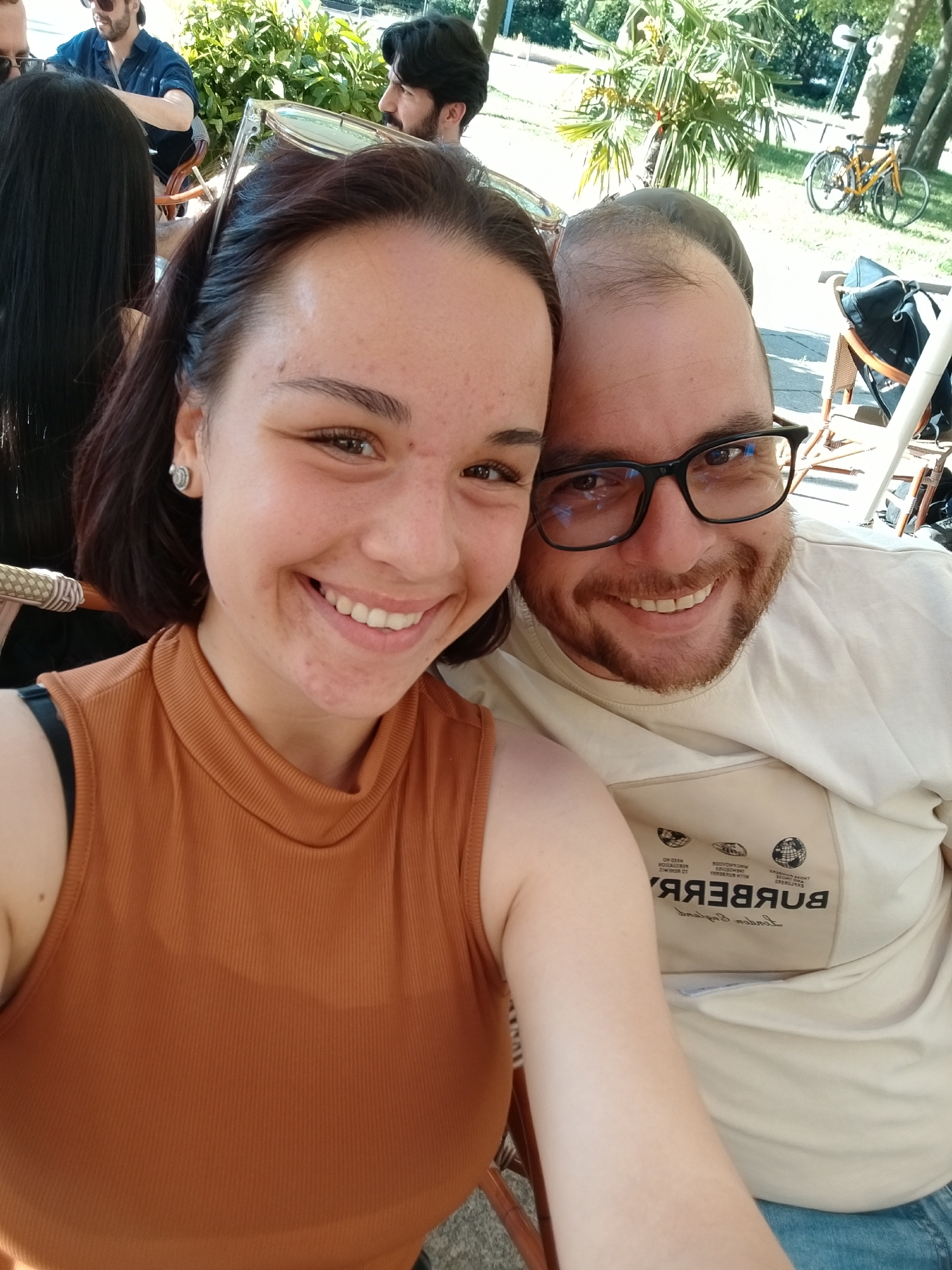 A selfie photograph shows a smiling woman and man seated outdoors in an unknown city. The woman has short brown hair, a brown top, and light skin. The man, who is right next to her, has dark hair, a beard, glasses, a light colored shirt with a "BURBERRY" inscription, and light skin. Both are looking directly at the camera and smiling, suggesting they are posing for the photo.
In the background, there are trees, sunlight, and other people. One of them is a man wearing glasses and a dark shirt. Another man with dark hair is visible as well. A yellow bicycle is parked in the background as well. The selfie appears to be taken in a park or a similar outdoor setting, possibly during a sunny day.