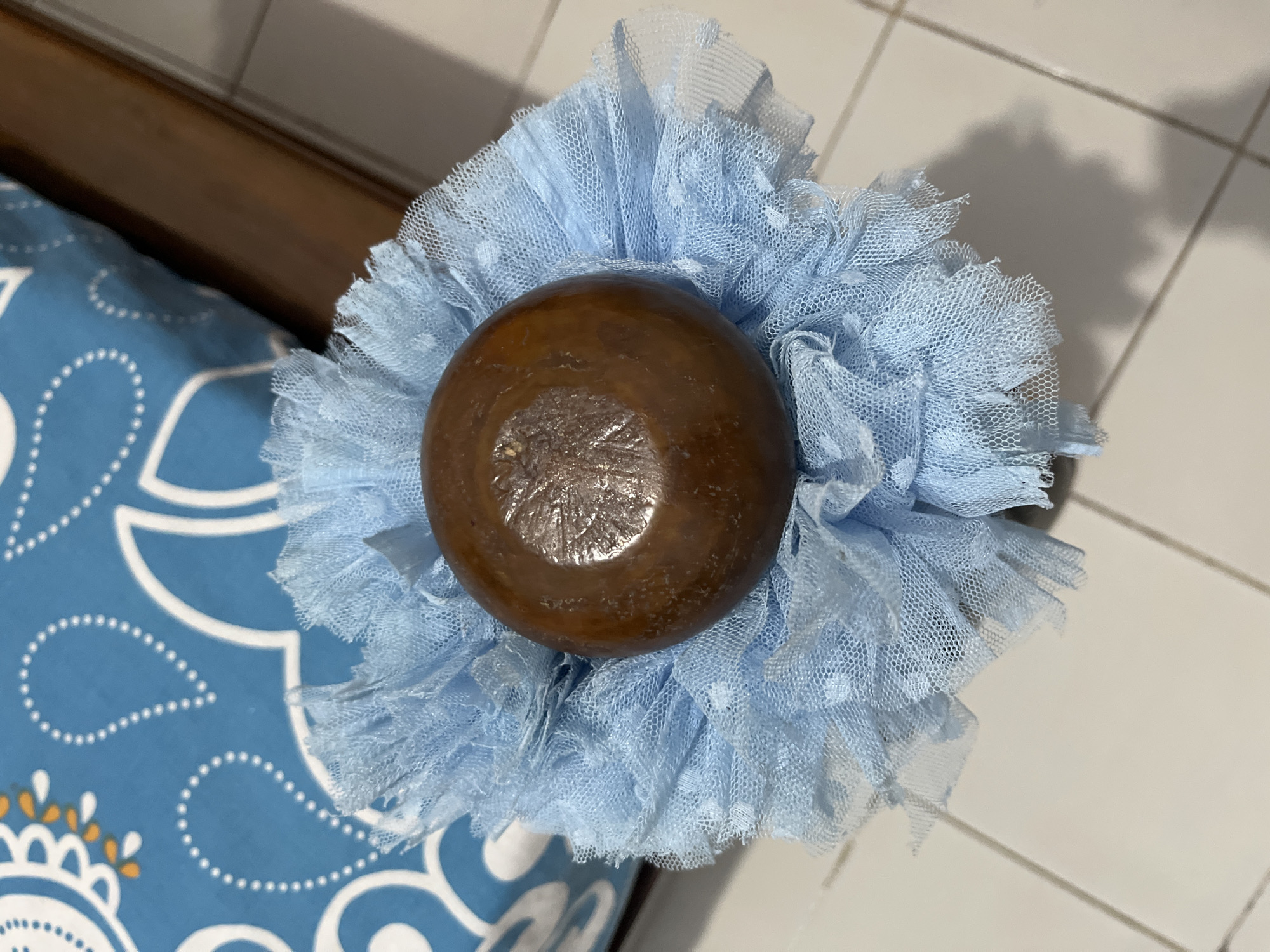 A close-up, overhead shot captures a brown, round object, possibly a wooden bead or stopper, nestled within a ruffled, pale blue net fabric. The fabric appears to be part of a decorative item, possibly a hair accessory or embellishment for clothing. The net has a subtle pattern of tiny white dots. In the background, a portion of a blue and white patterned textile, perhaps a pillow or blanket, is visible. The scene is set on a tiled surface, with grout lines indicating the flooring. The lighting casts a soft shadow to the lower right of the frame. The location is described as Glagah Kulon, Indonesia, though no specific urban features are identifiable in the image to confirm this.