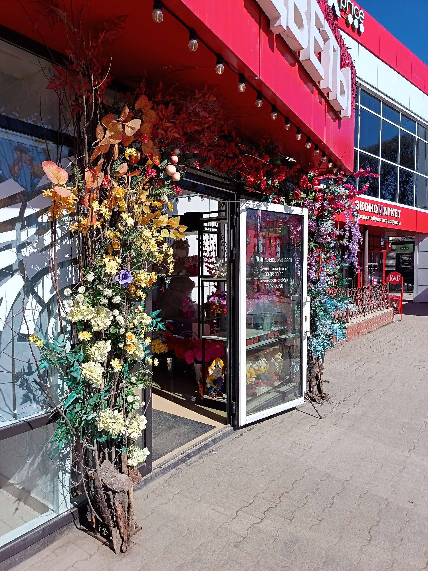 Here is a detailed description of the image:
**Scene:** The image captures the facade of a retail store, possibly a florist shop, in what appears to be a commercial area. The location is likely Osa, Russia, based on contextual clues such as the Cyrillic text on the storefront signage.
**Architecture and Storefront:** The building has a bright red awning and storefront. The store's name, "ЦВЕТЫ" (Flowers), is displayed in large, white Cyrillic letters on the facade. Underneath, there is text in Cyrillic, likely business hours and type of store, "ЭКОНОМАРКЕТ одежда обувь аксессуары". The building has glass windows that offer a glimpse into the interior. A secondary storefront bearing the logo "К&Б" is visible further down the street.
**Floral Decorations:** The entrance to the florist shop is adorned with an elaborate arrangement of artificial flowers and branches. The floral display includes a range of colors, from yellows and whites to oranges, reds, and purples. The flowers and branches are intertwined and create a decorative archway around the door.
**Entrance and Signage:** The store's entrance has glass doors with white frames. Signage with Cyrillic text is visible on the interior of the glass doors, likely indicating store hours or additional business information.
**Surroundings:** The store is located on a paved sidewalk. The overall environment appears to be urban and well-maintained.