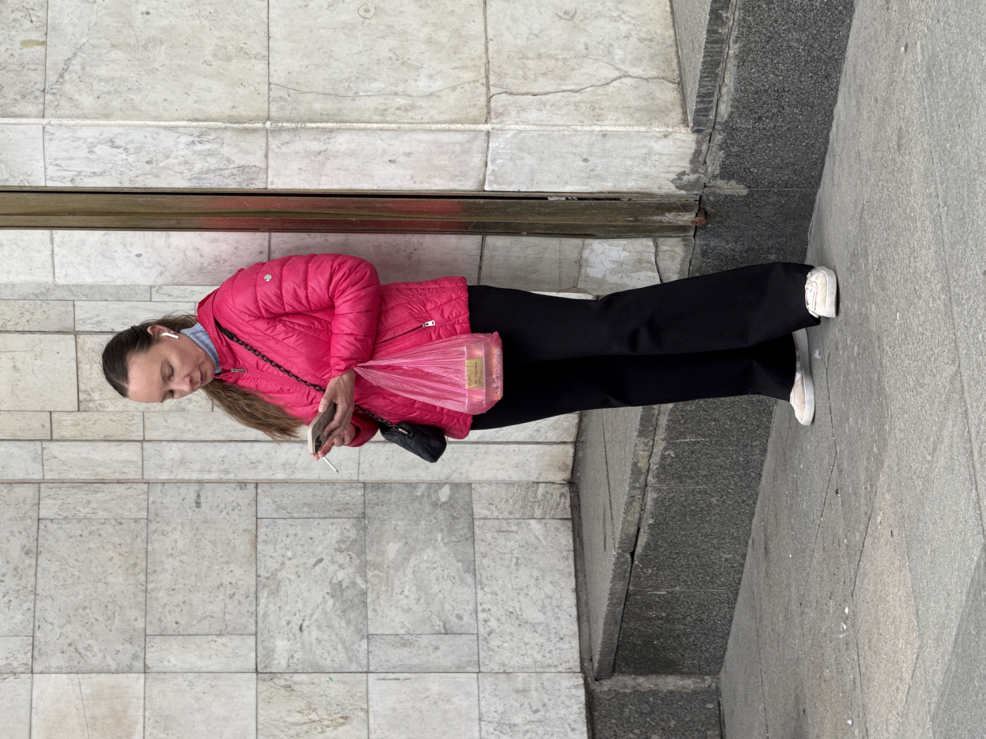 A woman stands on a sidewalk in Saint Petersburg, Russia. She has dark hair and wears an earbud. She is dressed in a bright pink puffer jacket and black pants, with white sneakers visible. She is holding a smartphone in one hand and a plastic bag filled with items in the other. A black purse hangs from her shoulder. She appears to be looking at her phone.
The background consists of a tiled wall and the gray sidewalk. The tile patterns suggest a public space. The scene is well-lit, indicating it is daytime. The photograph is taken from a slightly elevated angle.