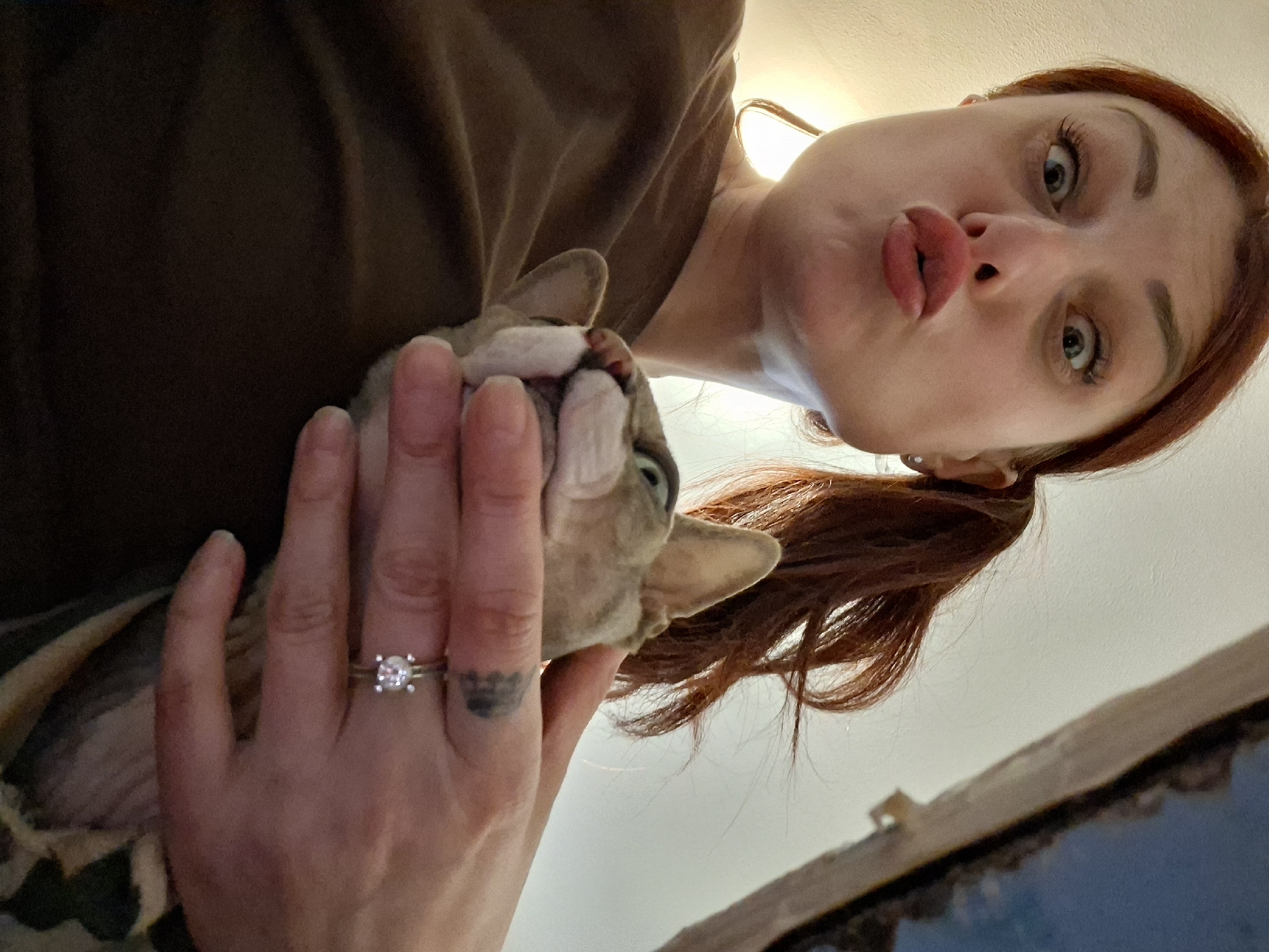 An adult female, characterized by reddish-brown hair styled in a ponytail, is captured from a low-angle perspective, making direct eye contact with the camera. Her facial expression features widened eyes and pursed lips. She wears a plain, dark brown garment. A silver-toned ring with a clear, prominent gemstone is present on her left ring finger, adjacent to a dark, patterned tattoo on the inner aspect of the same finger. She is holding a small, light-colored cat, consistent with a Sphynx or similar short-haired breed, cradled in her left arm and hand. The cat's head is visible, displaying green-yellow eyes and a slightly open mouth. The background consists predominantly of a light-colored wall or ceiling, illuminated by a warm light source from the upper left. A darker, structural element with a subtle blue tint is visible in the upper right background. This scene is presumed to be located in Забалоцкі сельскі Савет, Belarus.