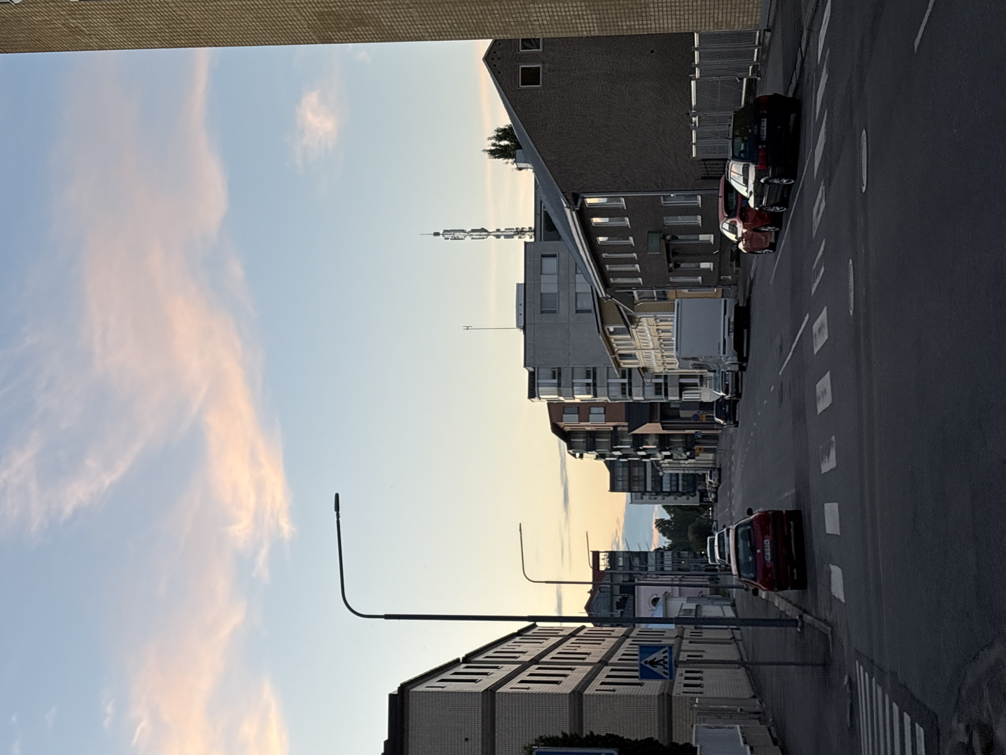 The image shows a street scene in Pori, Finland. The time of day appears to be either sunrise or sunset, with soft pink and orange hues in the sky and clouds. The street is lined with modern apartment buildings and some older, narrower buildings. There are several cars parked on the street and near the sidewalk, including a red Peugeot and a black Audi. A pedestrian crossing is visible in the foreground. A tall radio tower can be seen in the distance, above the buildings. The overall impression is of a quiet urban environment.
