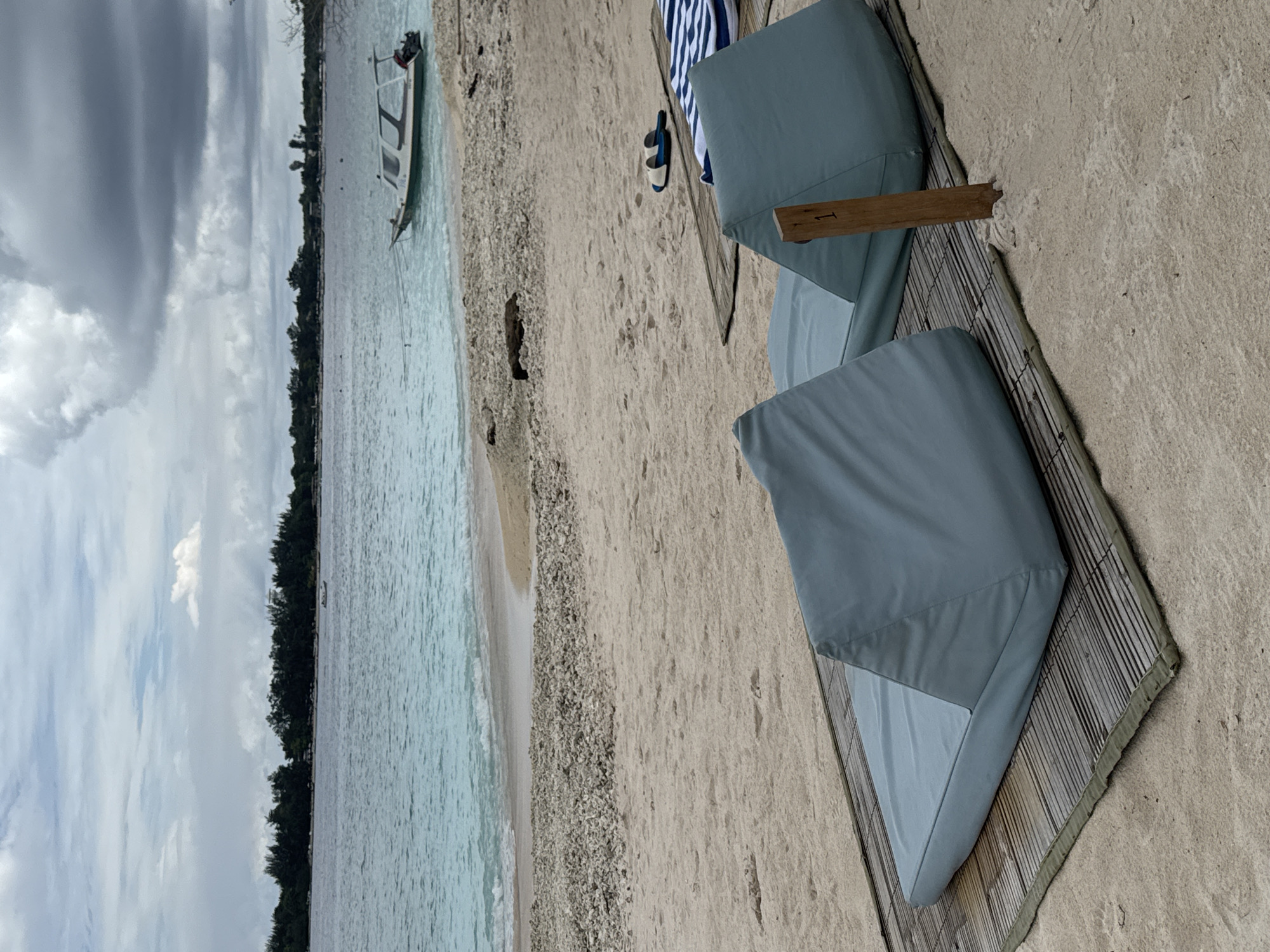 The image depicts a beach scene in an unknown location in Indonesia. The beach features white sand with sections of shell debris. Turquoise water laps against the shore, and a boat is visible in the distance. Lush green vegetation lines the coast. The sky is overcast with clouds.
In the foreground are two beach loungers, each with a blue cushion placed on a woven mat. A pair of sandals and a blue and white striped towel rest near one of the loungers. The overall impression is one of a relaxed and tranquil beach setting.
