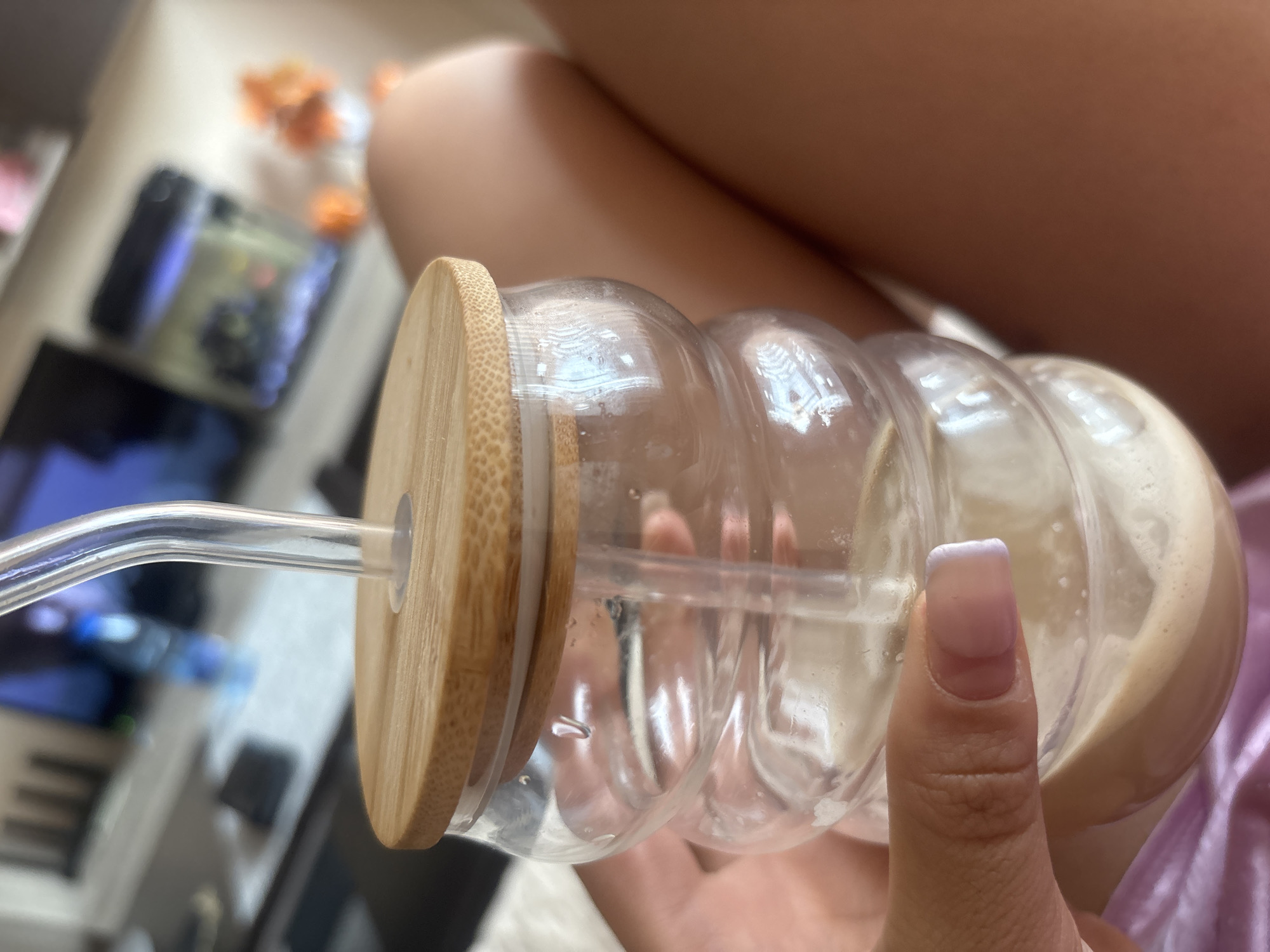 A clear glass beverage container with a bulbous, segmented shape and a light wooden lid, featuring a glass straw, is held in a person's hand. The beverage appears to be a light-colored liquid, possibly coffee or tea with milk. The person has a manicured fingernail with a French tip. The background is blurred, revealing a television screen and furniture. There is no information available about the location being in Plovdiv, Bulgaria.
