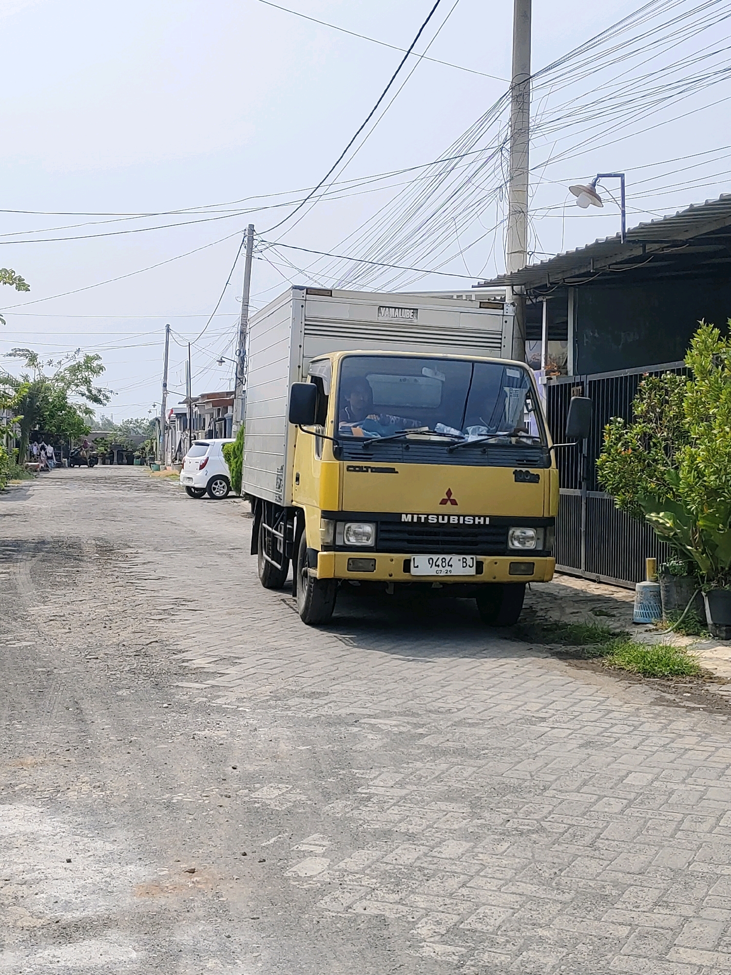 A yellow Mitsubishi Fuso truck with a white box body is parked on a paved street in a residential area. The truck has a license plate that reads "L 9484 BJ". A white car is parked on the side of the road behind the truck. There are houses and trees on both sides of the street. The sky is clear and blue. The image appears to be taken in Probolinggo, Indonesia.