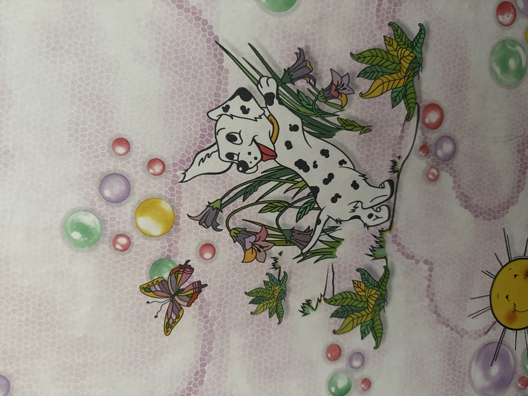 The image is a drawing of a Dalmatian dog playing in grass with flowers and a butterfly. The dog is white with black spots and is shown with its tongue out, appearing happy. The background is a light pink mosaic pattern with colorful bubbles scattered throughout. The drawing is whimsical and childlike, suggesting it might be part of a children's book or decoration. There is no identifiable location in the city of Bălți, Moldova, or any real-world setting depicted.