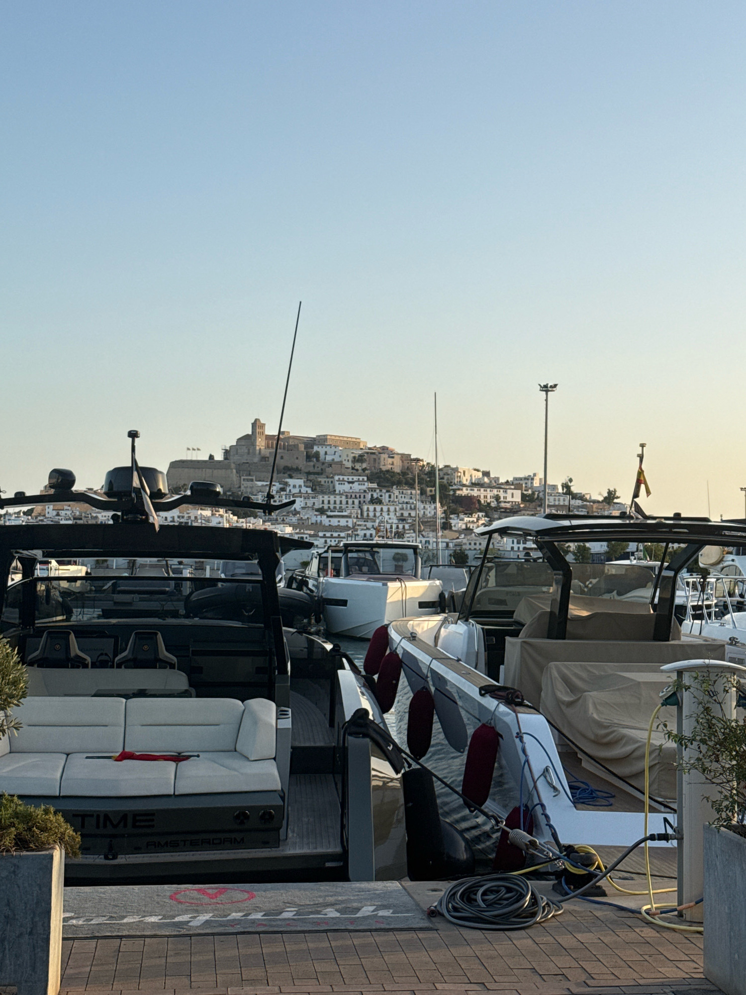 In the foreground, a black luxury yacht is docked. The yacht has white seating with a red item placed on it. The rear of the yacht displays the text "TIME AMSTERDAM". Also visible are mooring lines, fenders, and a yellow electrical cable connected to the boat. In the middle ground, other boats are docked, and a white structure with a tower and lights is present. In the background, the historic town of Eivissa, Spain, is visible on a hill, characterized by white buildings and a prominent fortress. The sky is clear and light blue, indicating daylight.