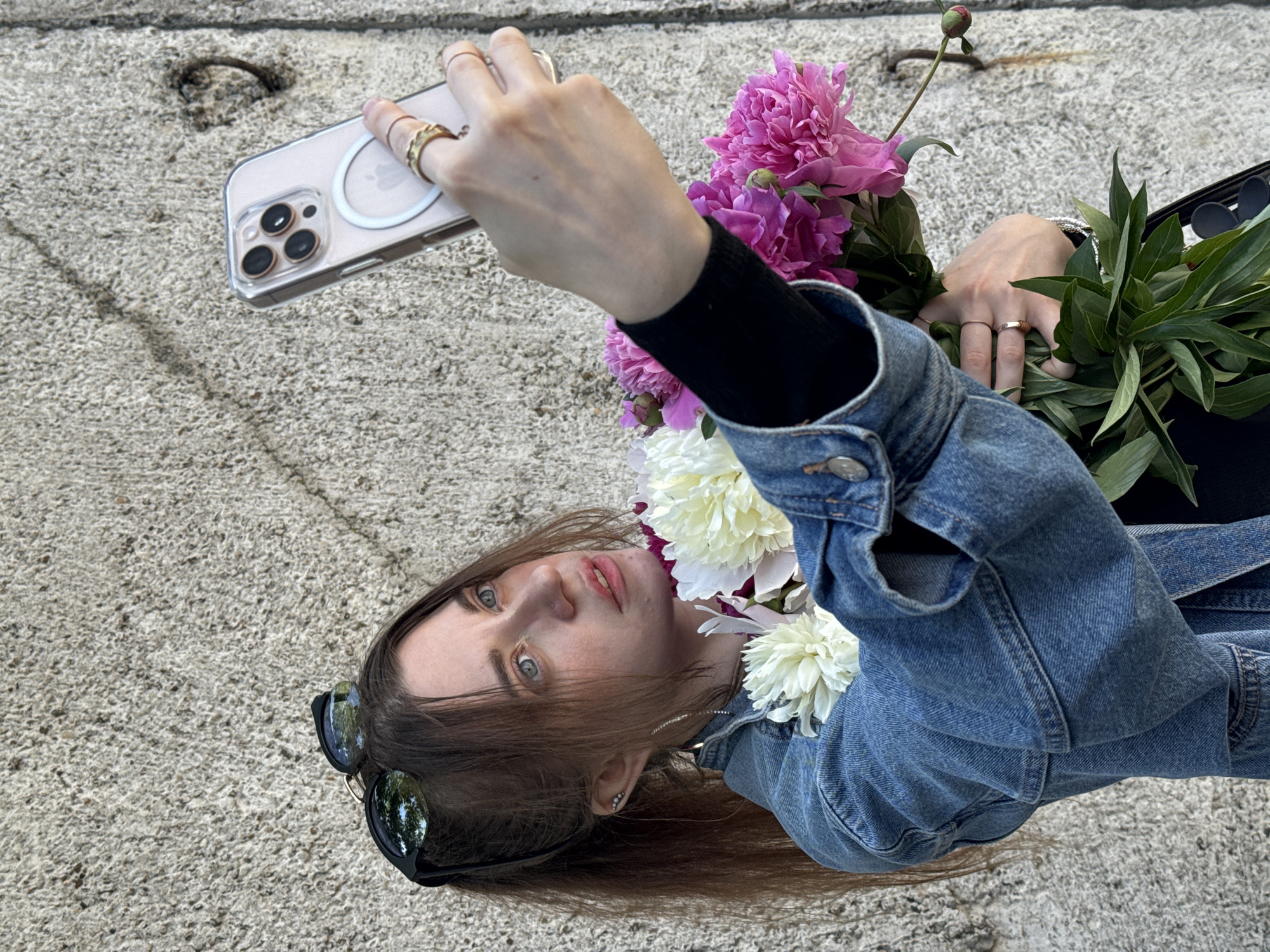 A woman with brown hair pulled into a ponytail is taking a selfie with a bouquet of pink and white peonies. She is wearing a denim jacket over a black top and sunglasses are perched on her head. She is also wearing rings on her fingers and a bracelet. The background is a rough, concrete wall. The image appears to have been taken outdoors on a sunny day. The location is the city of Chaikovsky, Russia.