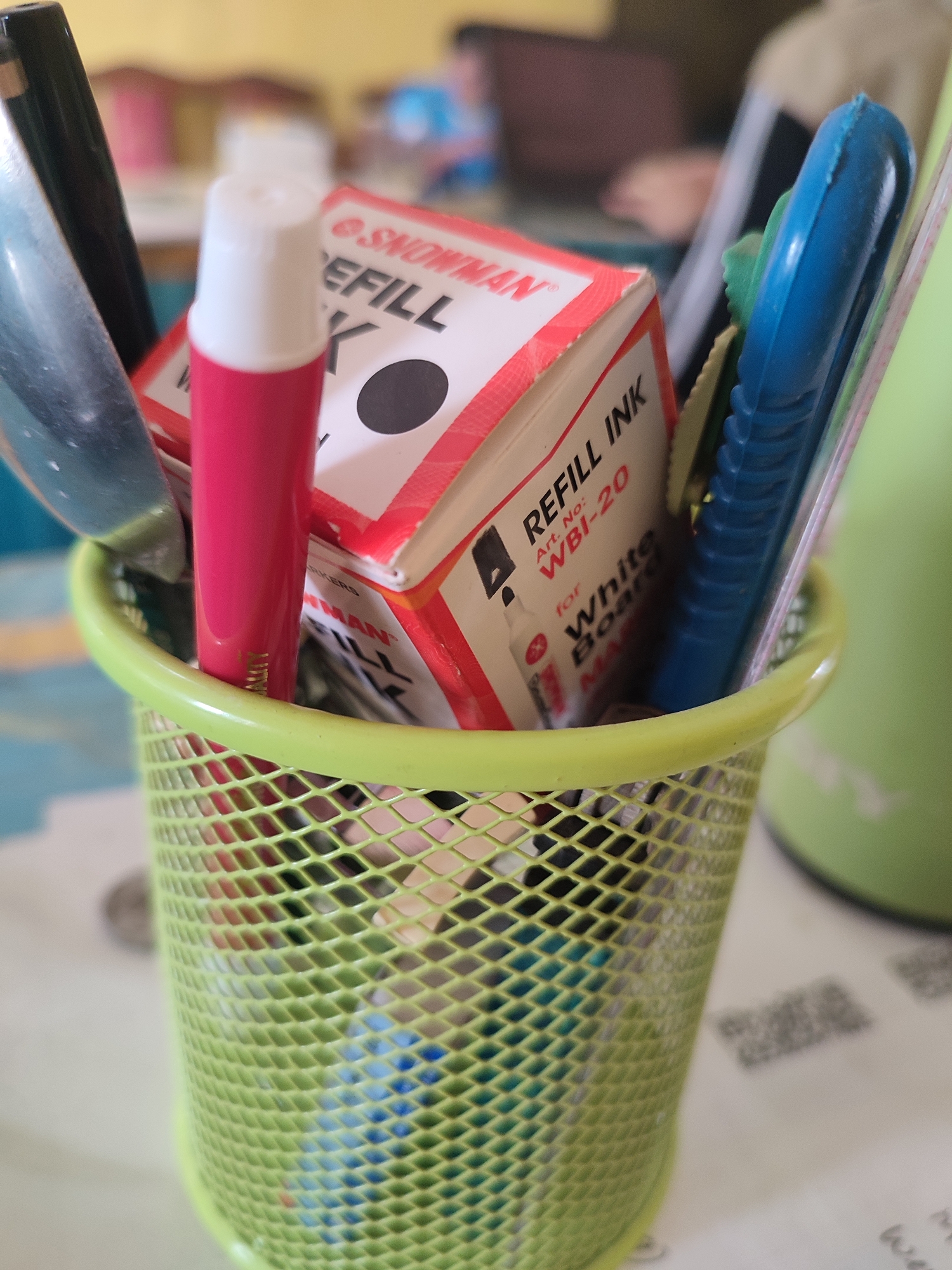 An overhead shot shows a light green, mesh pen holder filled with office supplies. A black marker, a silver letter opener, a pink tube with a white cap, and a box labeled "SNOWMAN REFILL INK WBI-20" are visible. A blue utility knife with a green grip is also present, along with other blurred items. The pen holder sits on a surface that appears to be a table, with papers and a blurred green object in the background. The lighting suggests an indoor setting. There are no people visible in the image, and no specific location in Nusa Tenggara, Indonesia can be determined from the provided visual information.