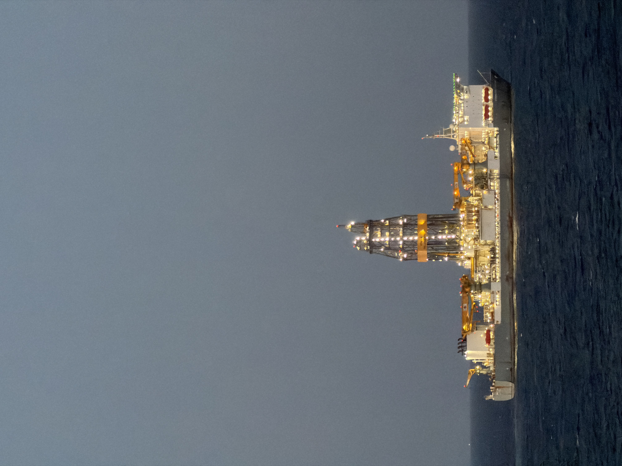 The image depicts a large, brightly lit vessel at sea, seemingly an offshore drilling platform or a specialized construction ship. The vessel is positioned on the right side of the frame, contrasting against a dark, smooth body of water. Above, the sky is a uniform, dark grey, suggesting either night or twilight conditions. The vessel itself is complex, with multiple levels of platforms, cranes, and support structures illuminated with bright lights. The overall scene conveys a sense of industrial activity taking place in an isolated marine environment. The location is Unknown, Unknown.
