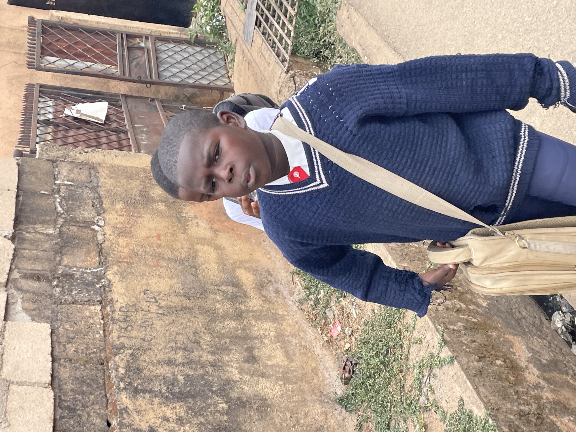 A medium shot, slightly rotated clockwise, captures a young person standing outdoors, looking towards the camera with a neutral expression. The main subject, a child or teenager with short dark hair, wears a navy blue V-neck sweater with white and thin blue stripes along the neckline, and similar striped cuffs. A white collared shirt is visible underneath, adorned with a small red pin or badge on the left side of the chest. They are also wearing dark blue trousers.
A light beige cross-body bag or satchel with a wide strap is slung over their left shoulder, and their right hand lightly grasps the bag's handle near their hip.
In the background, to the left of the subject, is a weathered, light brown building with two prominent windows fitted with decorative metal bars and security grilles. The wall appears made of brick or stone with a rough plaster finish. Below the windows, the wall transitions to a lower, rough-hewn stone foundation. Faint, dark, illegible writing or graffiti is visible on the wall to the right of the subject's head in the mid-ground.
The ground features a mix of dirt, sparse green vegetation, and what appears to be a concrete or paved path to the right, consistent with an outdoor street or pathway setting. The lighting suggests it is daytime. The overall scene appears to be in an urban or suburban area of Jos, Nigeria.