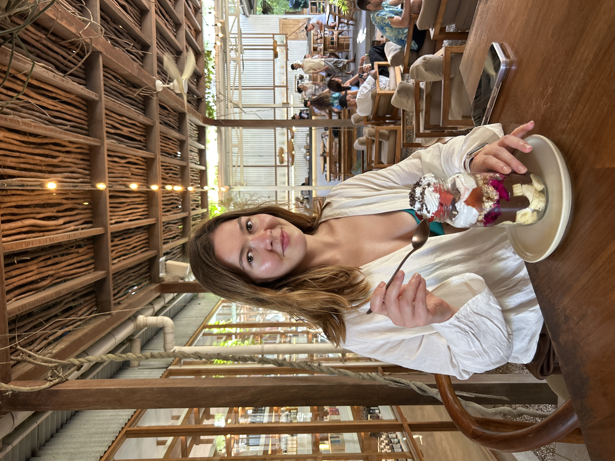 In Kuta Selatan, Indonesia, a woman sits at a wooden table in an open-air cafe with a thatched roof and exposed wooden beams. She wears a white open-fronted shirt over a blue top and holds a spoon over a large dessert in a glass jar. The dessert contains layers of chocolate, fruit, granola, and whipped cream topped with chocolate shavings and a piece of fruit. A smartphone rests on the table next to her. Other patrons are visible in the background, seated at tables and walking around the cafe, which features wooden furniture and shelves stocked with items. The overall ambiance is casual and tropical.