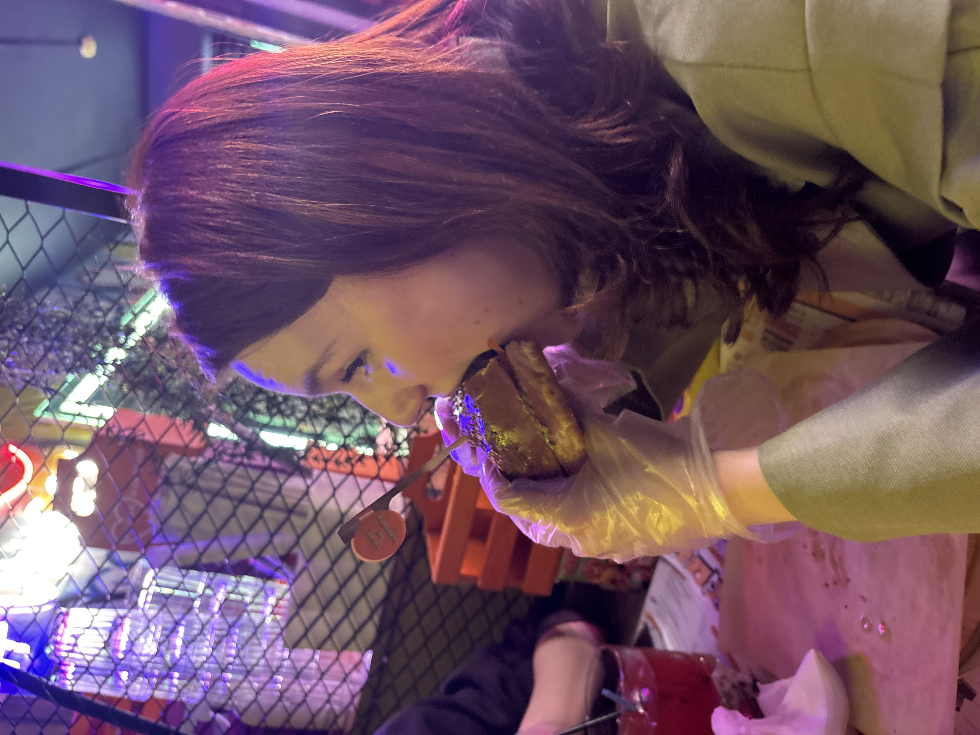 A woman with brown hair is biting into a sandwich while wearing clear plastic gloves. A metal cage is visible in the background. Part of an orange object is visible near her face. In the foreground, there is a red cup and a white napkin.