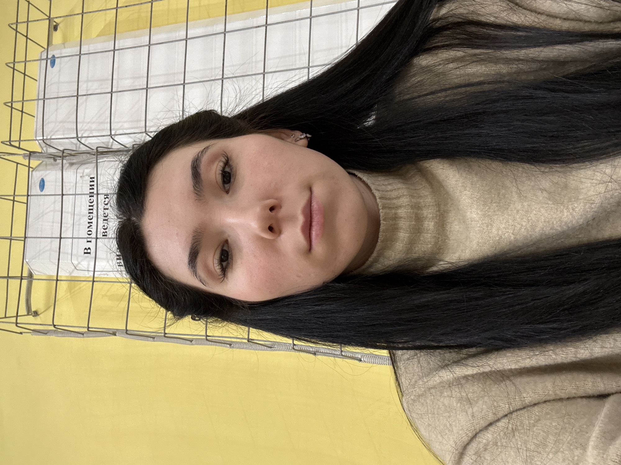 A selfie taken by a young woman with dark hair and light skin, wearing a beige sweater. The background consists of a yellow wall, a wire mesh, and white boxes with text in Russian, suggesting the location may be in Izhevsk, Russia.
