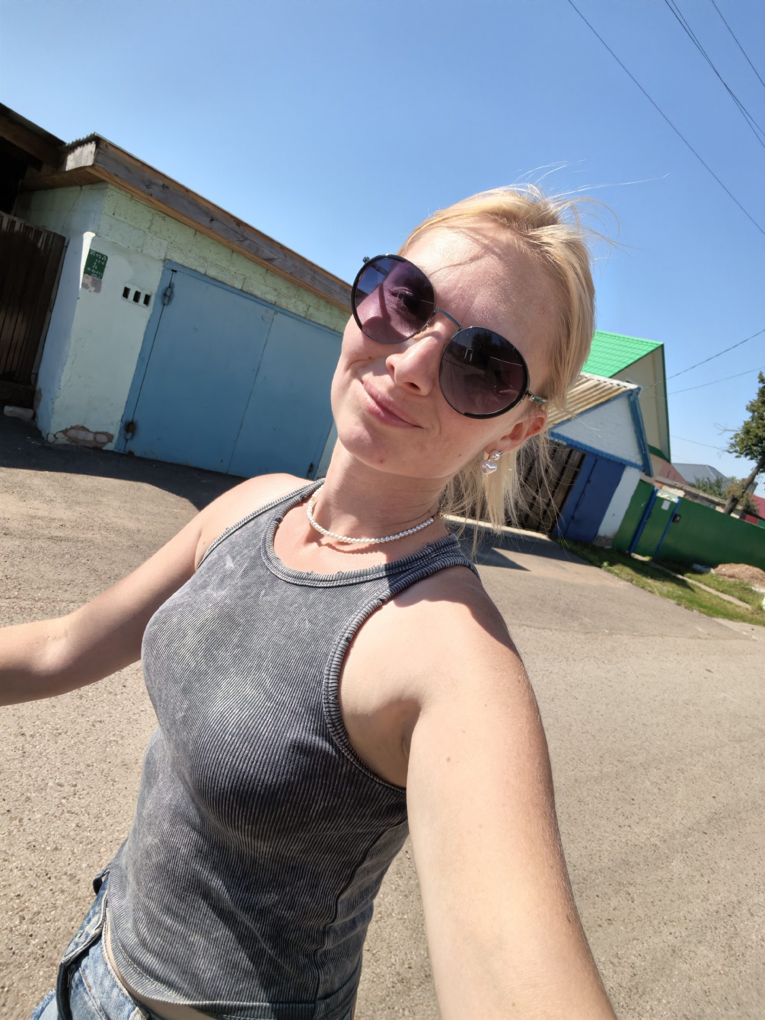 A blonde woman wearing sunglasses, a grey tank top, and a pearl necklace smiles at the camera while taking a selfie outdoors. The background shows a light blue garage with a wooden roof, a green-roofed building with a blue door, and a clear blue sky. The setting appears to be a residential area with a paved road in the foreground. The woman is wearing denim shorts, which are partially visible. The image is taken on a sunny day.