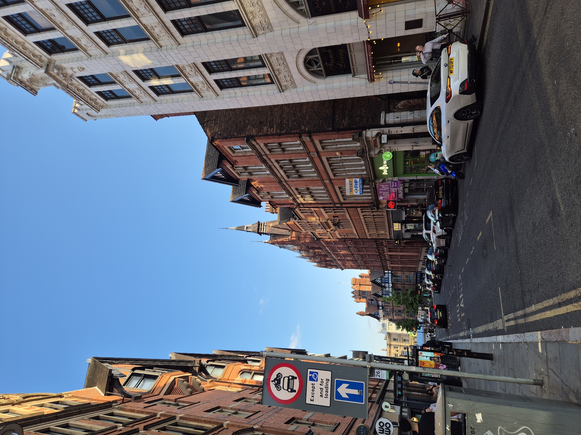 The image captures a street scene in Nottingham, United Kingdom, likely during late afternoon or early evening given the lighting. The street is lined with a mix of red brick and white stone buildings, exhibiting varied architectural styles. On the left, a tall brick building with multiple windows dominates the foreground, with a traffic sign displaying restrictions for motorcycles and cars, permitting exceptions for disabled parking and loading. Further down the street, other brick buildings with ornate facades and steeply pitched roofs are visible. On the right, a white stone building with detailed carvings and arched windows stands prominently.
The street itself is paved with asphalt and has visible double yellow lines indicating parking restrictions. Several cars are parked along the curbs and on the street. A white BMW is parked on the right, facing away from the camera, with its license plate clearly visible. Other cars, including black sedans and SUVs, are also present. Pedestrians are seen on the sidewalks, some walking, others standing or interacting. A woman in a grey sweatshirt stands on the right sidewalk, and another is near the entrance of a building. A man in a black shirt is seen on the left sidewalk. Various shops and businesses are identifiable along the street, indicated by signage such as "French Living," "Nail Art," and "Panda Tea." A trash bin is situated on the left sidewalk. The sky is clear and blue, with a hint of clouds.