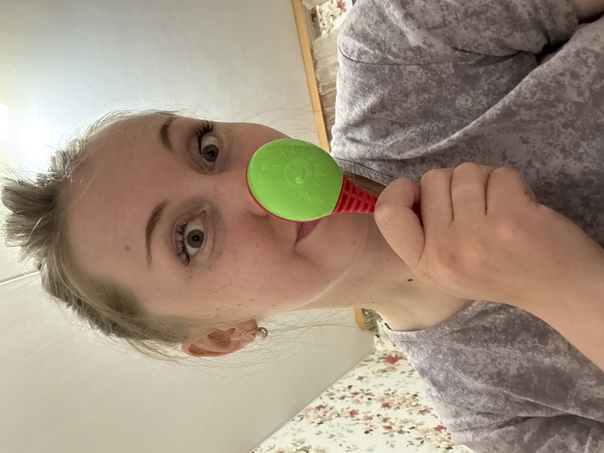 A woman is holding a green and red maraca. She is wearing a light purple patterned shirt. Her hair is blonde. The location is Unknown, Unknown. The background includes a white wall and a floral patterned surface. The woman is looking directly at the camera.