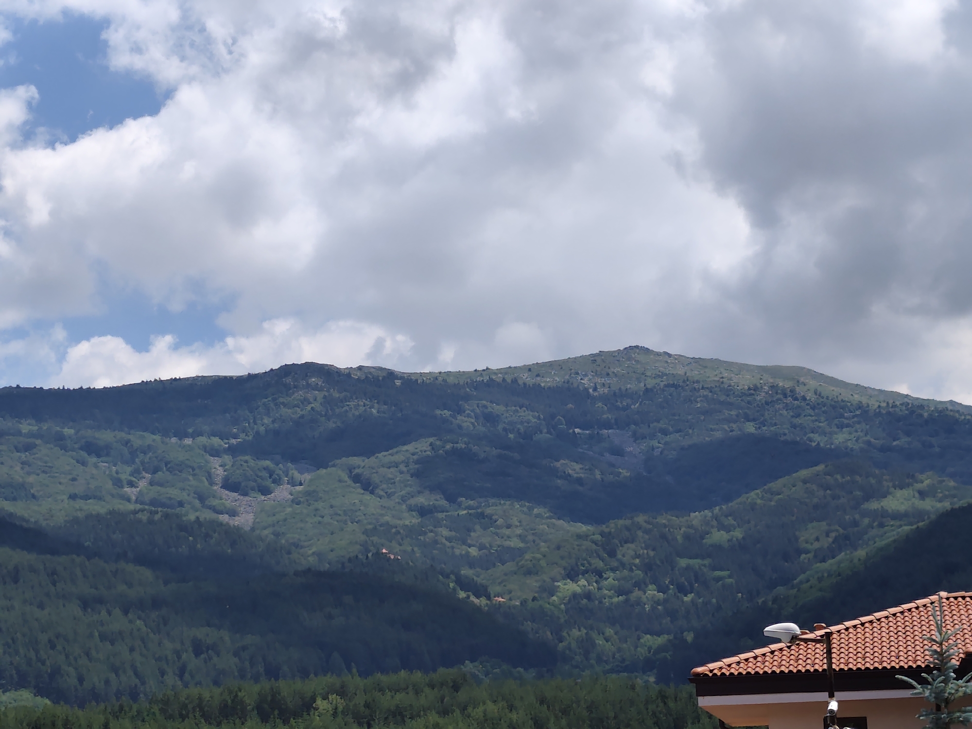 The image captures a vast mountainous landscape under a cloudy sky, with a residential building in the foreground. The mountains, covered in dense green vegetation and some rocky patches, rise majestically, their slopes disappearing into the distance. The sky is dramatic, with large, billowing white and grey clouds partially obscuring the sun. In the lower right corner of the frame, the terracotta-tiled roof of a building is visible, along with a street lamp and a small evergreen tree. The scene appears to be set in Kladnitsa, Bulgaria, a location known for its natural beauty and mountainous terrain. The overall impression is one of natural grandeur and tranquil residential life integrated into the landscape.