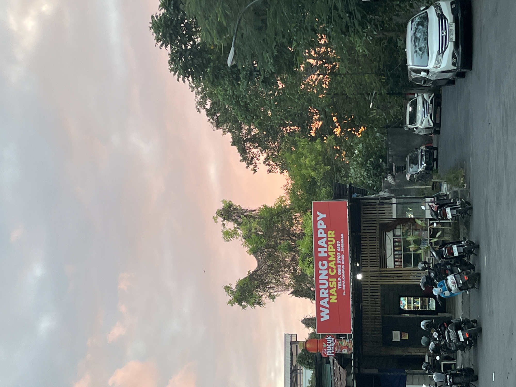 The image captures a street scene in Kuta Selatan, Indonesia, during what appears to be sunset or early evening. The sky is a gradient of soft pinks and blues, indicative of twilight. In the foreground, several motorcycles are parked on the asphalt. To the left, a small roadside food stall, "WARUNG HAPPY NASI CAMPUR," is visible, with a red sign displaying its name and contact information. A bottle of "Teh Pucuk Harum" is placed near the sign. The stall itself has a bamboo-like facade and appears to be open for business. Further down the street, a few cars are parked or driving, including a white SUV in the immediate right foreground. Lush green trees line the right side of the street, with some of their branches visible against the colorful sky. A street lamp is mounted on a pole amidst the foliage. The overall atmosphere suggests a casual, everyday moment in a semi-urban environment.