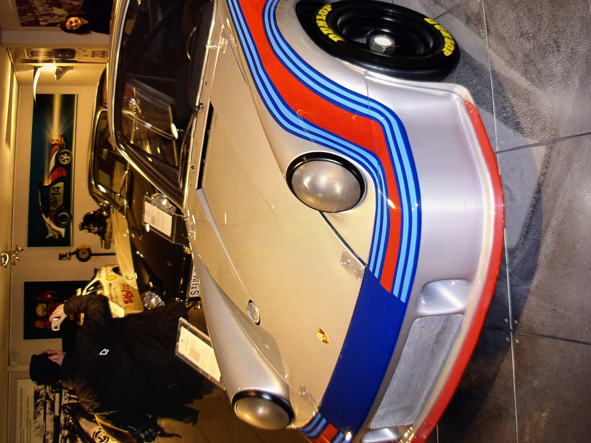 The image shows a silver Porsche race car with red, white, and blue racing stripes on display indoors. A man in a dark jacket is standing behind the car, holding a camera and looking at the car. A woman is visible in the background, smiling. The car is the main subject, with its front end prominently featured. The environment appears to be a showroom or museum, with other cars and display items visible in the background. The flooring is tiled. The scene appears to be in Kyiv, Ukraine, as suggested by the context.