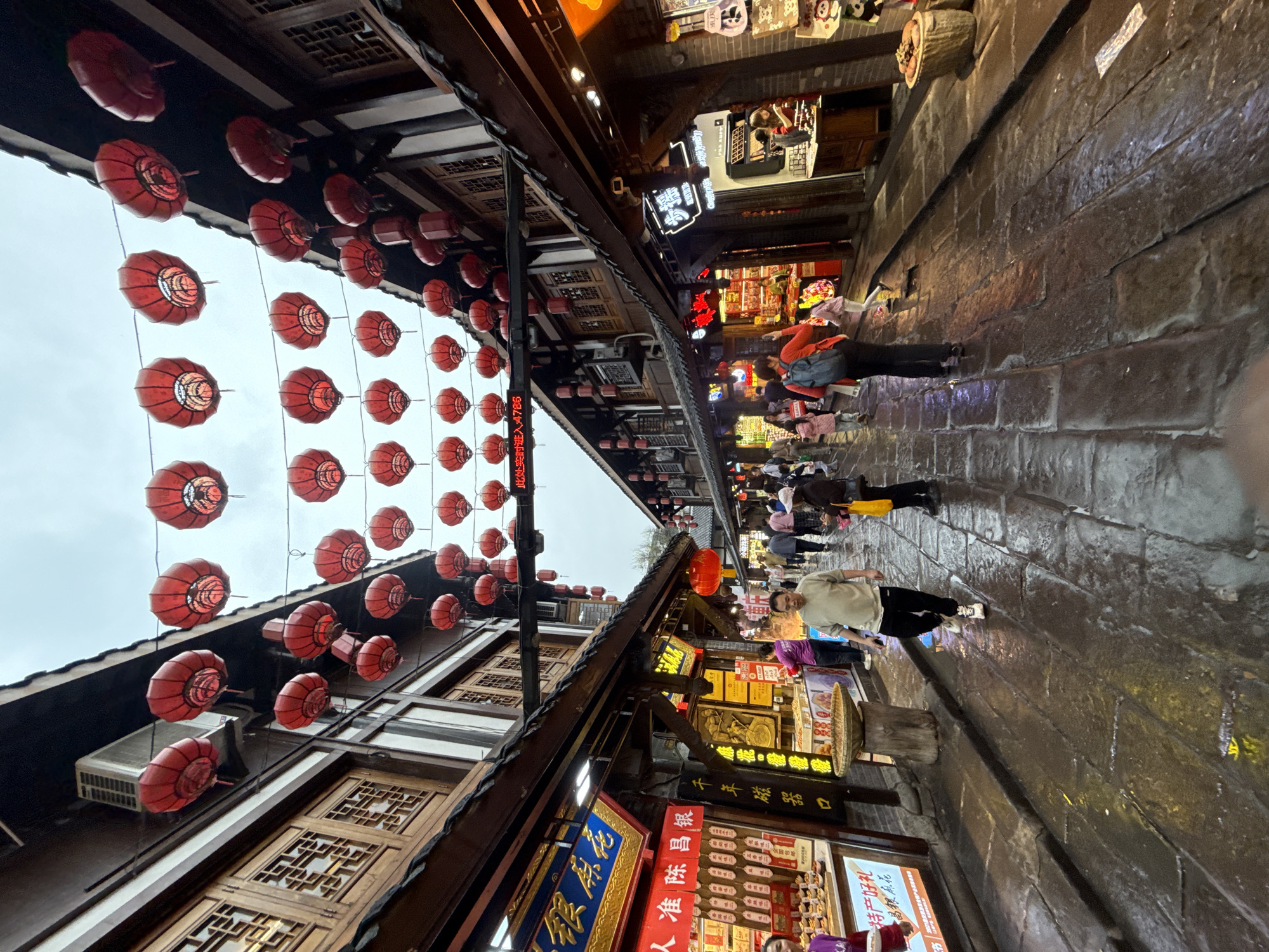 The image shows a street in the Shapingba District of Chongqing, China. Red lanterns are strung above the street, which is lined with shops featuring traditional Chinese architectural details. People are walking along the wet, cobblestone street. Several signs are visible, including those advertising "Chen Changyin" and "Millennial Porcelain Mouth." The image captures the vibrant atmosphere of the street, with its blend of traditional architecture and commercial activity.