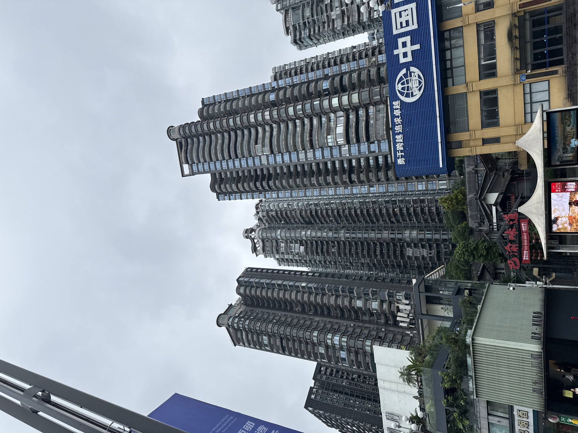 The image shows a street-level view in Nan'an District, China, featuring tall, modern residential buildings with distinctive wavy balconies. The buildings are predominantly gray and feature numerous windows. Commercial signage in Chinese is visible, including a large blue sign with the characters "中国" and a logo that suggests a financial institution. A yellow building is seen beside the blue sign. Street-level businesses and signs advertising alcohol are present, partially obscured by the viewpoint's angle. The sky is overcast.