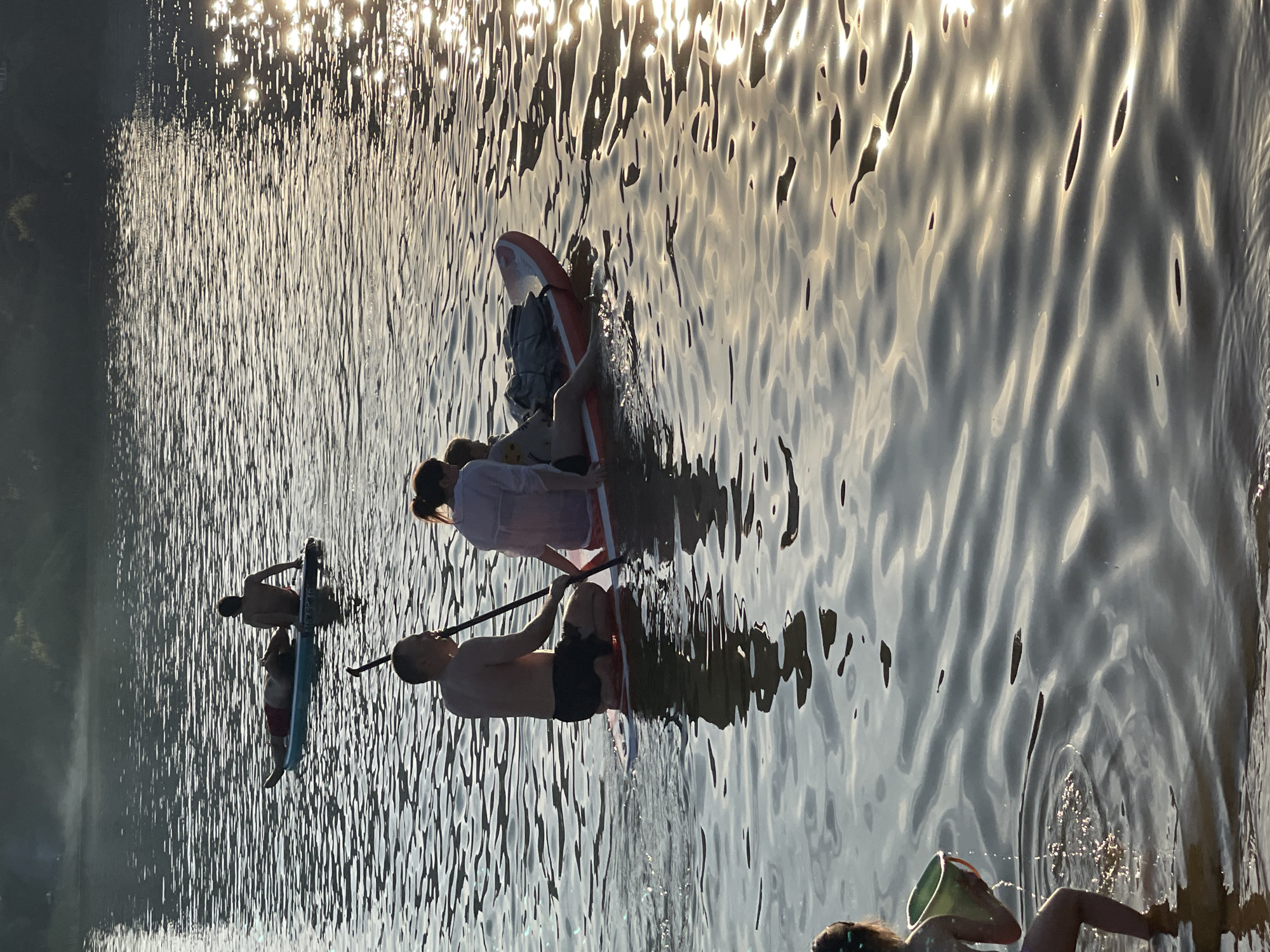 The image depicts an outdoor recreational scene on a body of water located in Moscow, Russia, characterized by low-angle sunlight reflecting intensely on the water surface from the right, creating significant glare and sparkling effects. The background features a dark, indistinct landmass, likely a treeline, under a hazy sky.
In the mid-ground, a shirtless man in dark shorts kneels on an orange and grey paddleboard, holding a black paddle, facing left. Behind him, a woman in a light-colored, sheer cover-up sits, her back mostly to the viewer. A child, dressed in a light top and dark shorts, sits or lies further forward on the same paddleboard, facing right, with a grey fabric item nearby.
Further in the background, two individuals are visible on a separate teal and blue paddleboard. One person, shirtless and wearing red swim shorts, is lying prone on the board. The second person is sitting upright at the rear, also shirtless, with both individuals generally facing left.
In the foreground, a young child is standing in shallow water, partially submerged. The child is holding a light green bucket, from which water is actively spilling, creating ripples and splashes in the immediate vicinity.