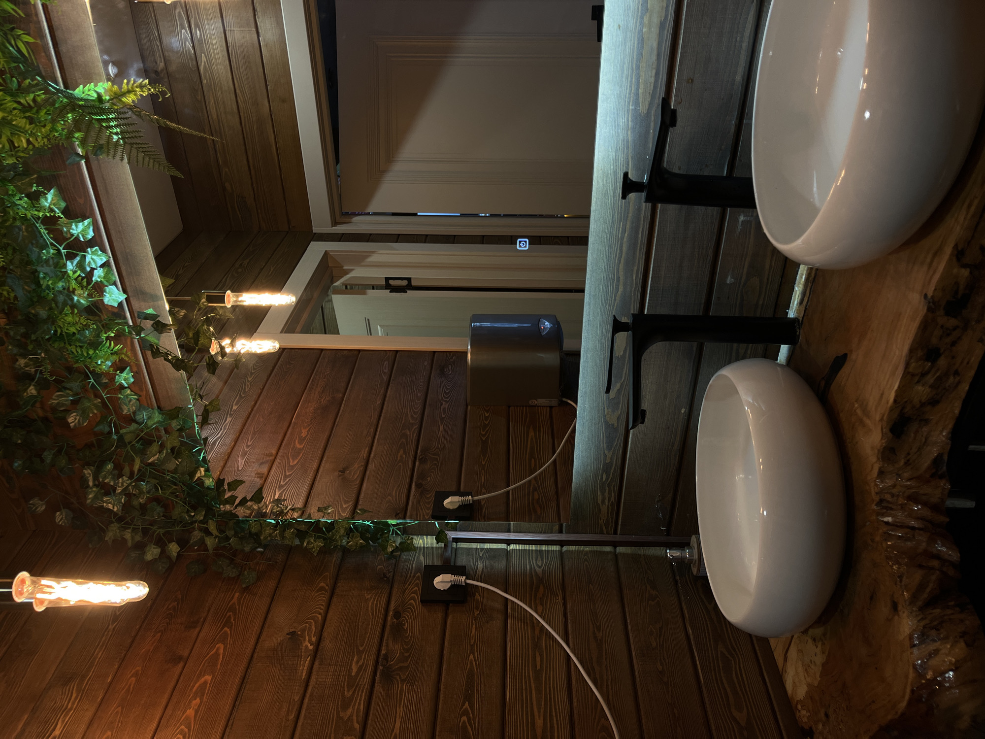 Here's a detailed description of the image:
**Setting:** The image appears to be an interior shot of a restroom, possibly in a commercial establishment. The walls are covered in horizontal wood paneling, and there is faux greenery (ivy and ferns) decorating the left side of the wall.
**Objects:**
* Two white, bowl-shaped sinks are visible on the right side of the frame. They are mounted on dark, wrought-iron style brackets.
* A wooden countertop or vanity supports the sinks.
* Two light fixtures are attached to the wall, emitting a warm, incandescent glow.
* A hand dryer is mounted on the wall between the sinks.
* Electrical cords and outlets are visible.
* A partially visible doorway suggests access to another area.
**Environment:** The lighting is warm, creating an ambiance. The combination of wood paneling and greenery gives the restroom a rustic or nature-inspired feel.
**Inferences:** The location in Neftekamsk, Russia, is not visually verifiable from this image alone.