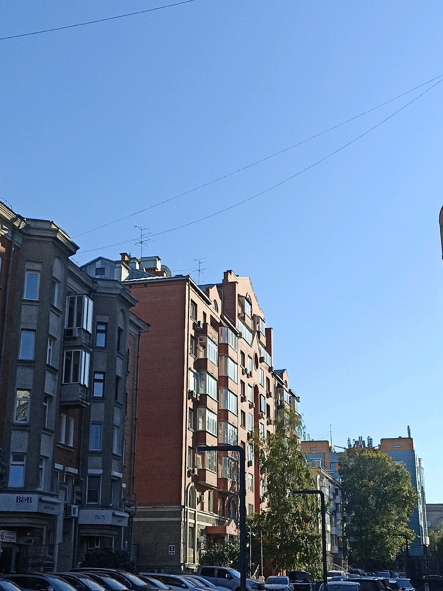 A low-angle, full shot captures a street scene in Novosibirsk, Russia, under a clear blue sky. The foreground is dominated by a row of parked cars, facing left. Behind them, a line of multi-story buildings runs across the frame. The building on the far left is grey with visible window frames and some projecting balconies. Next to it is a brick building with many windows, some of which are balconies. To the right, a taller building with blue and grey facades and orange accents is partially visible. Trees with green foliage are interspersed between the buildings. The overall impression is of a typical urban street with residential and commercial structures.