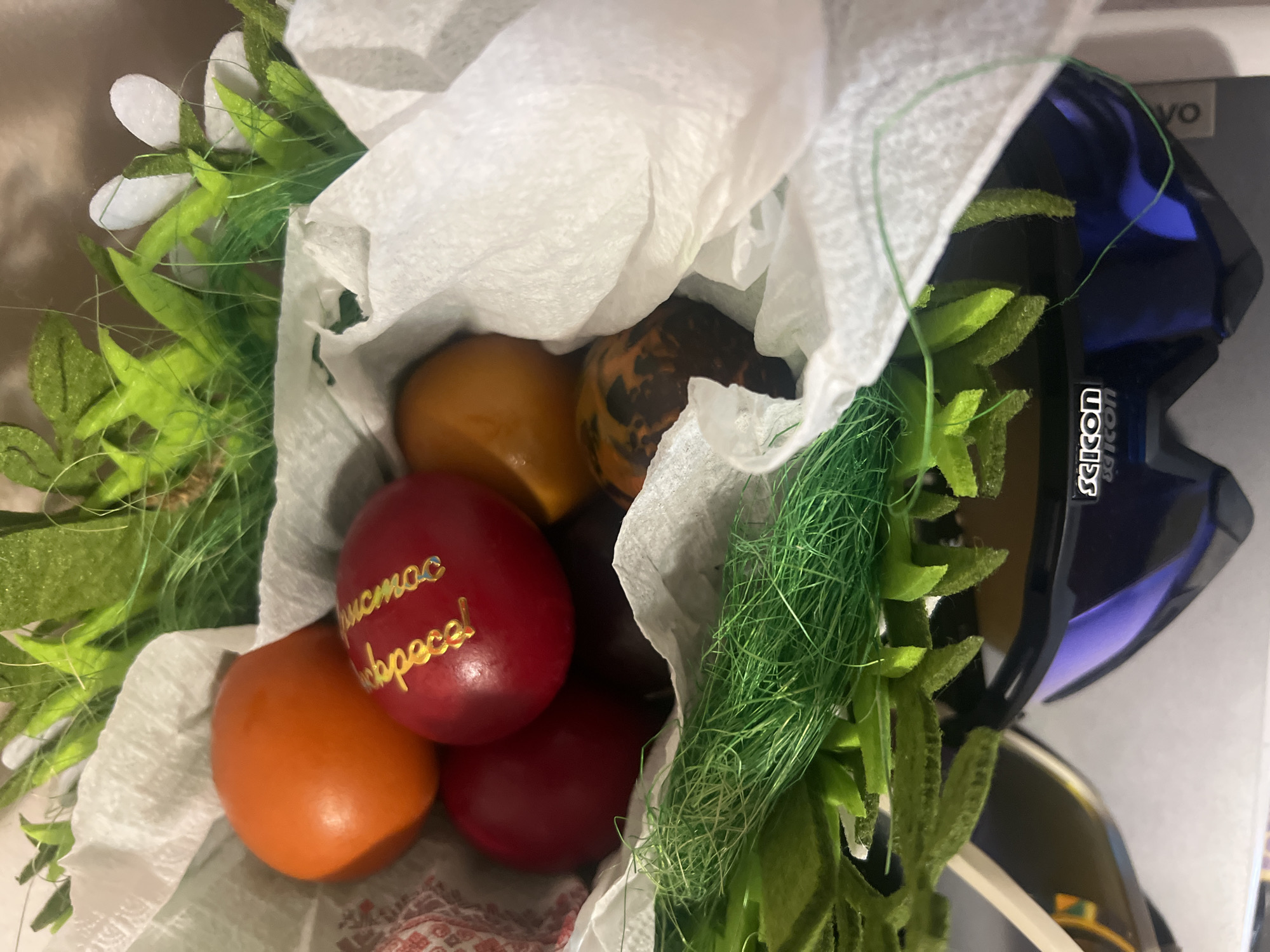 A close-up shot shows a collection of colorful Easter eggs nestled within white tissue paper and surrounded by artificial greenery. The eggs are various shades of red, orange, and brown, with one red egg bearing a golden inscription. The tissue paper is crumpled, adding texture to the arrangement. Artificial green stems and leaves protrude from the sides, framing the eggs. In the background, partially visible, is a dark blue object labeled "Sicon". Given the presence of Easter eggs and the style of decoration, this likely represents a small Easter basket or display. The location within Plovdiv, Bulgaria is not discernible from the image alone.