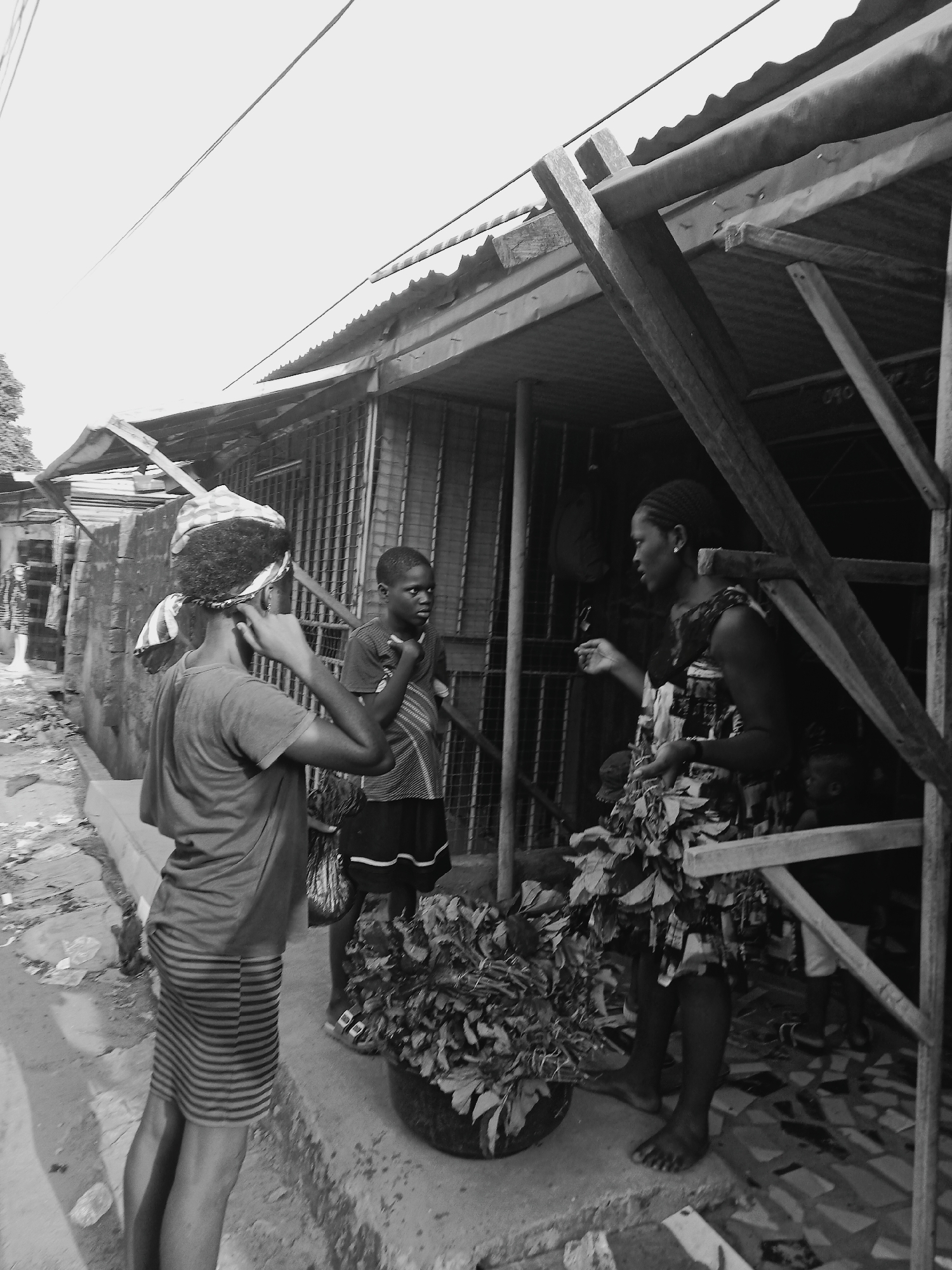 A black and white photograph depicts three individuals in what appears to be a street market setting in Onitsha, Nigeria. The location is suggested by the architectural style and overall atmosphere.
A woman in a patterned dress stands under the roof of a building and interacts with two younger people outside. The woman under the roof has dark skin and is barefoot. She is looking at the other two people.
The other two people are standing outside the building. The person closest to the woman under the roof is also dark skinned, wearing shorts and a striped shirt, and looking at the woman. The third person in the photo has their back to the camera and is wearing a patterned scarf/hair covering, a striped skirt, and a darker t-shirt. This third person has their hands on their head.
In front of the people, there is a large pot that appears to be filled with plants or leafy vegetables. The scene is framed by the building's wooden supports. The environment is cluttered, with what looks like waste or debris on the ground. The buildings are simple, and the entire image conveys a sense of a bustling but impoverished urban area. The sky is light, either naturally or perhaps from photo overexposure.