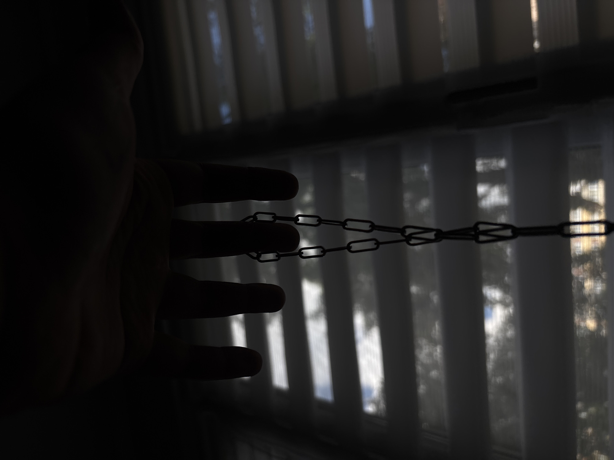 The image depicts a close-up view of several fingers, silhouetted against a backdrop of horizontal blinds. A metal chain hangs from between the fingers, with its links clearly visible. The lighting is dim, with light filtering through the blinds, creating horizontal stripes of illumination and shadow. The background suggests an indoor environment, possibly a room with a window covered by blinds. There is no information within the image that can be used to determine its location in Saint Petersburg, Russia.