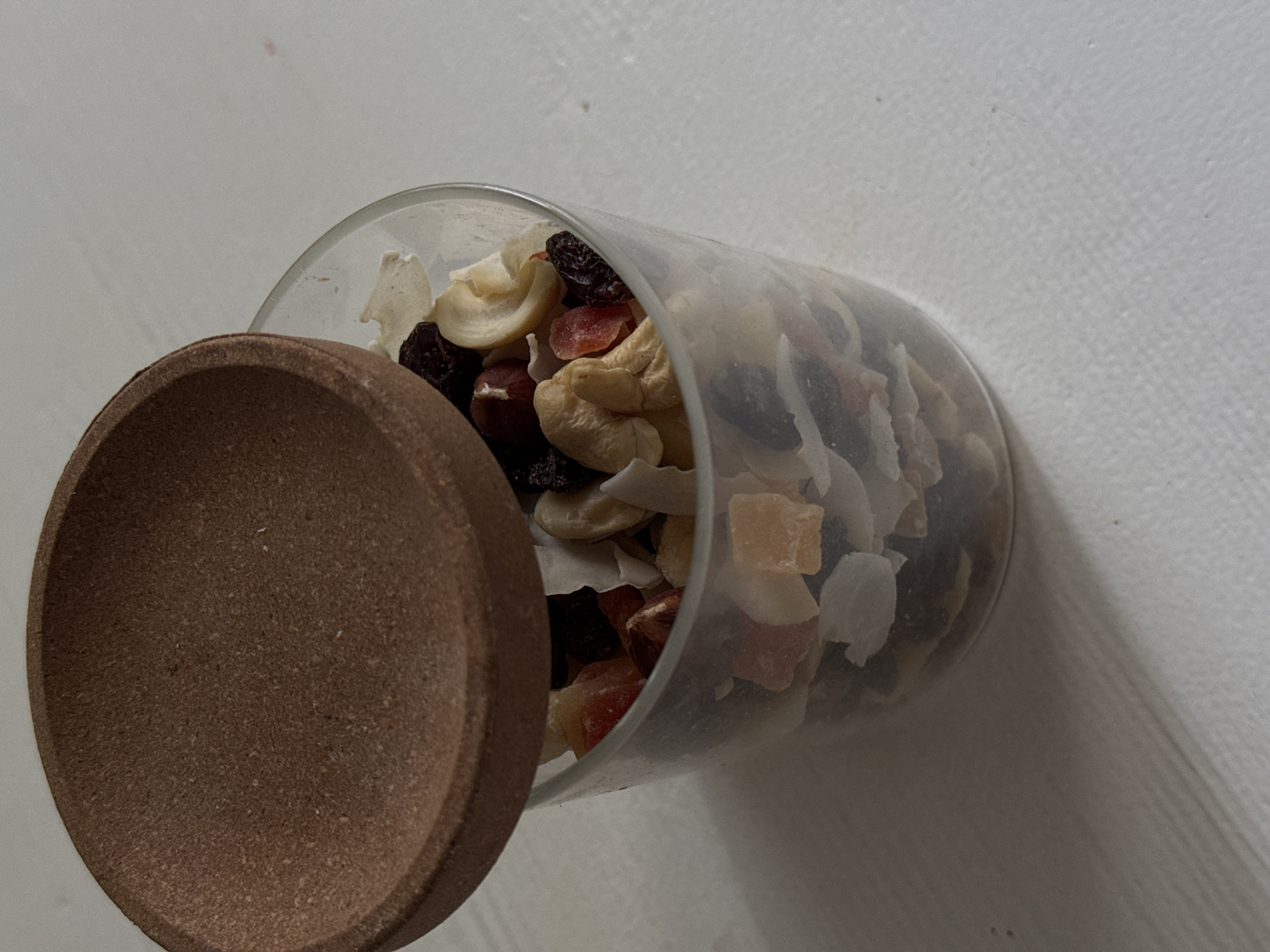A close-up, high-angle shot shows a clear glass jar filled with trail mix. The trail mix contains various ingredients including raisins, cashews, dried fruit pieces (such as papaya and possibly mango), and coconut flakes. A round, brown lid made of cork or a similar material rests partially over the top of the jar, angled to the left. The jar and its contents are situated on a white surface, possibly a table, with a textured grain. The lighting creates shadows on the surface, indicating the light source is from the right. There is no discernible location in Siegburg, Germany indicated in the image.