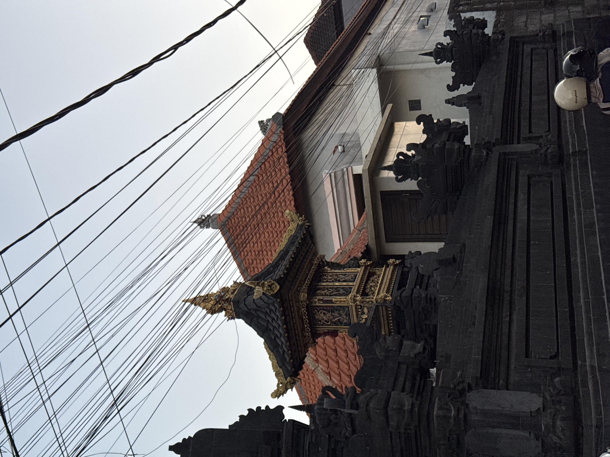 This image captures a view of a traditional Balinese gate and building, likely located in Denpasar, Indonesia, with a prominent array of tangled overhead electrical wires against a pale sky. The gate is constructed from dark stone, intricately carved with decorative elements that suggest religious or cultural significance. A smaller, ornate structure with a golden tiered roof sits atop a section of the gate. Behind the gate, a building with a terracotta tiled roof is visible, featuring a light-colored facade and a dark-framed window. The scene is dominated by the dense network of electrical wires crisscrossing the frame, a common sight in many urban and suburban areas of Indonesia. Two individuals wearing helmets are partially visible in the foreground, suggesting they are on a motorcycle, their presence adding a contemporary element to the otherwise traditional architectural context. The overall impression is one of a blend of old and new, with the intricate Balinese design juxtaposed against the modern infrastructure of electrical lines and the presence of motorized transport.