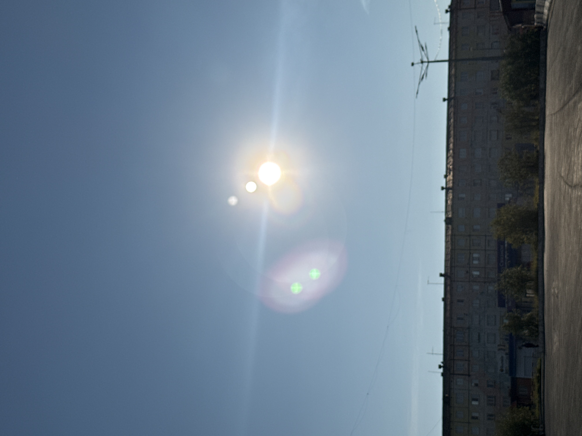 The image depicts a bright, sunny day in Norilsk, Russia, with a clear blue sky dominating the upper portion of the frame. The sun, appearing as a large, bright orb, is visible slightly to the right of center, accompanied by lens flares and a smaller, fainter orb above and to its left. On the left side of the sky, there are two distinct green circular lens flares.
Below the sky, a long, multi-story apartment building stretches across the frame. The building appears to be constructed of concrete or similar materials, with numerous windows. The facade shows signs of wear and tear, consistent with an older structure. Antennas are visible on the roof of the building.
In front of the apartment building, a line of trees and bushes are sparsely planted. The ground in the foreground is a dark, paved surface, possibly a parking lot or road, with faint tire marks visible. To the right of the apartment building, a distinctive street lamp with multiple arms is present. The overall impression is of an urban environment in a northern latitude.